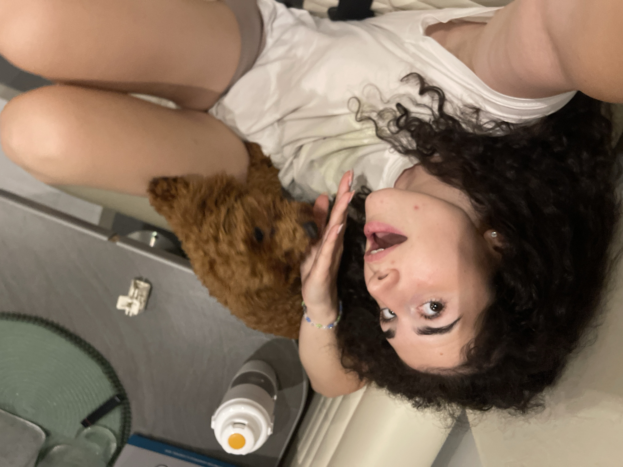 The image depicts a young woman and a small dog in an indoor setting, specifically in Koprinka, Bulgaria. The woman is lying on her back, angled so her head is towards the lower center of the frame, looking directly upwards at the camera with her mouth slightly agape and eyes wide. Her dark, curly hair is spread around her head. She wears a white t-shirt. A multi-colored beaded bracelet is visible on her left wrist, and a small stud earring is present in her right ear. A small red blemish is visible on her chin. Her left hand is raised, with fingers slightly curled, positioned near her face. A small, brown, fluffy dog, appearing to be a poodle, is partially visible near the woman's upper thighs/hip area, its head looking downwards and away from the camera. To the left of the woman (from her perspective), a grey flat surface, possibly a table or desk, holds a white and grey water bottle with an orange button, a dark cylindrical object (possibly a pen or vape), and a small, irregularly shaped white object resembling an electronic component. A portion of a green woven mat is also visible on this surface. The immediate foreground shows a light-colored surface, likely furniture.