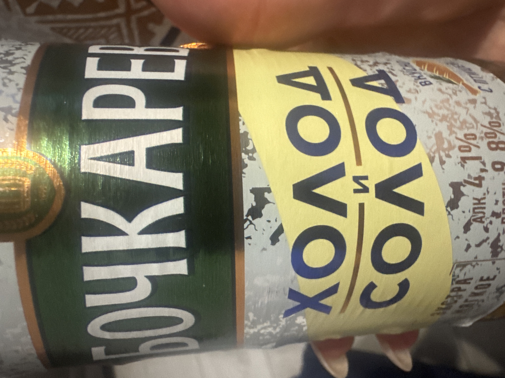 The image shows a close-up of a beverage can. The can has a green band at the top with white Cyrillic lettering that appears to read "БОЧКАРЕВ". Below the green band is a yellow section with larger, dark blue Cyrillic lettering. The top line reads "ХОЛОД" and the second line reads "СОЛОД". There is also text on a white label at the bottom, including "4,1%" and "9,8%". The can is held by a hand with manicured nails. The background is blurry and appears to be an indoor setting. There is no indication of the city of Mariupol, Ukraine, in the image.