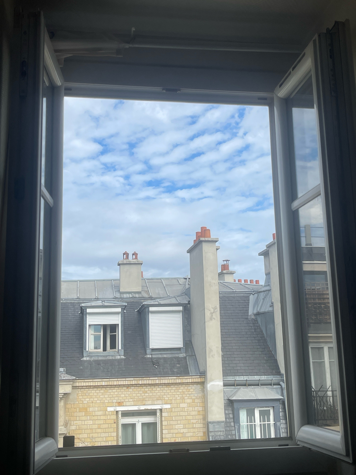 The image shows a view from an open window looking out onto Parisian rooftops under a partly cloudy sky. The architecture suggests a classic Haussmannian style, common in Paris. The rooftops are predominantly covered in grey slate, with multiple chimneys, some topped with orange clay pots. Several windows with white roller shutters are visible, some partially retracted. A beige brick building forms the lower layer of the view, with a window featuring white shutters and a balcony with black railings. The interior of the room from which the photo is taken frames the view, with white window frames and a white ceiling visible. The scene captures an ordinary urban landscape in Paris, France, on a day with diffused light filtering through the clouds. There are no people or animals present in the scene.