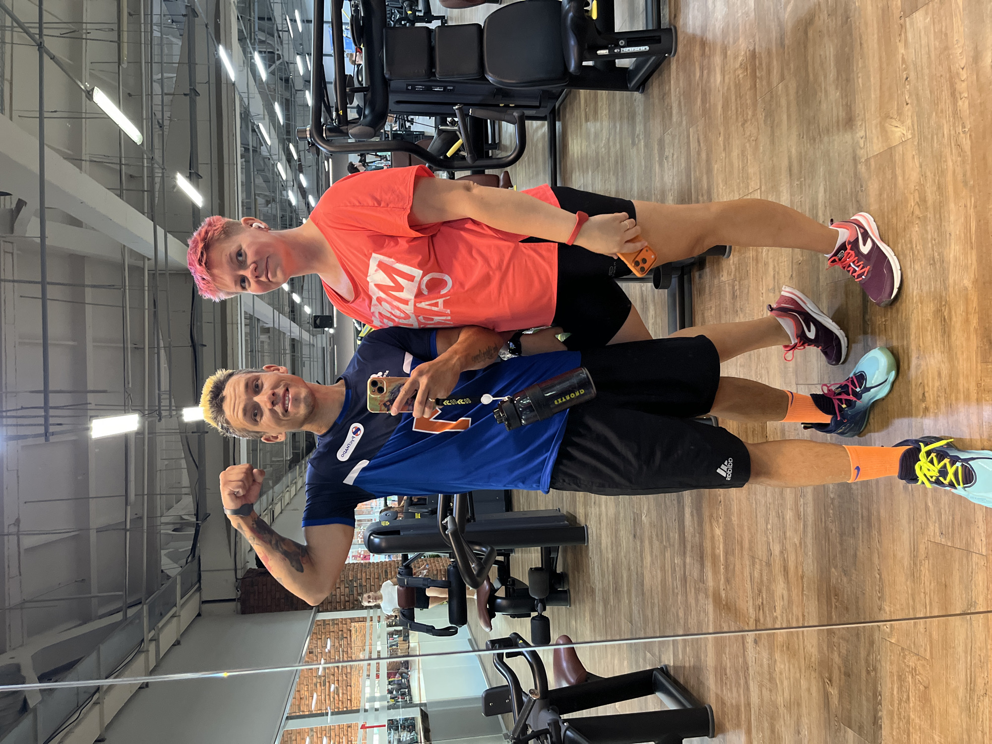 This is a full-body, mirror selfie taken in a gym. The image features a man and a woman, both dressed in athletic wear. The man on the left has bleached blonde hair and is wearing a blue and white t-shirt, black shorts, and colorful athletic shoes. He is flexing his bicep for the camera and holding a smartphone. The woman on the right has short, pink hair and is wearing a coral-colored t-shirt, black shorts, and dark athletic shoes. She is looking towards the camera with a neutral expression. In the background, gym equipment, including weight machines and benches, are visible. The flooring is wood, and the walls appear to be brick in some areas. The setting is consistent with a gym environment, potentially located in Chaikovsky, Russia, given the provided context.