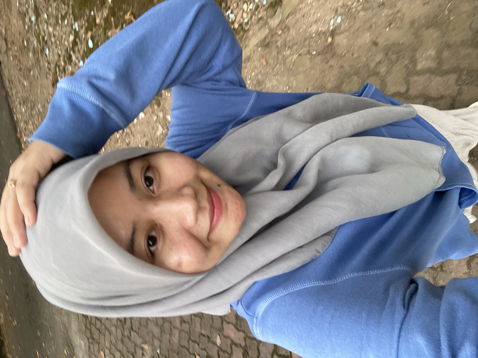 A young woman, wearing a light blue sweatshirt and a grey hijab, is taking a selfie outdoors. She is standing on a paved walkway with a reddish-brown cobblestone pattern. The ground to her right is dirt and scattered with small, light-colored objects. Her left hand is raised, touching the top of her head, and her right arm is bent. She is smiling at the camera. The background is blurred and appears to be a natural outdoor setting.