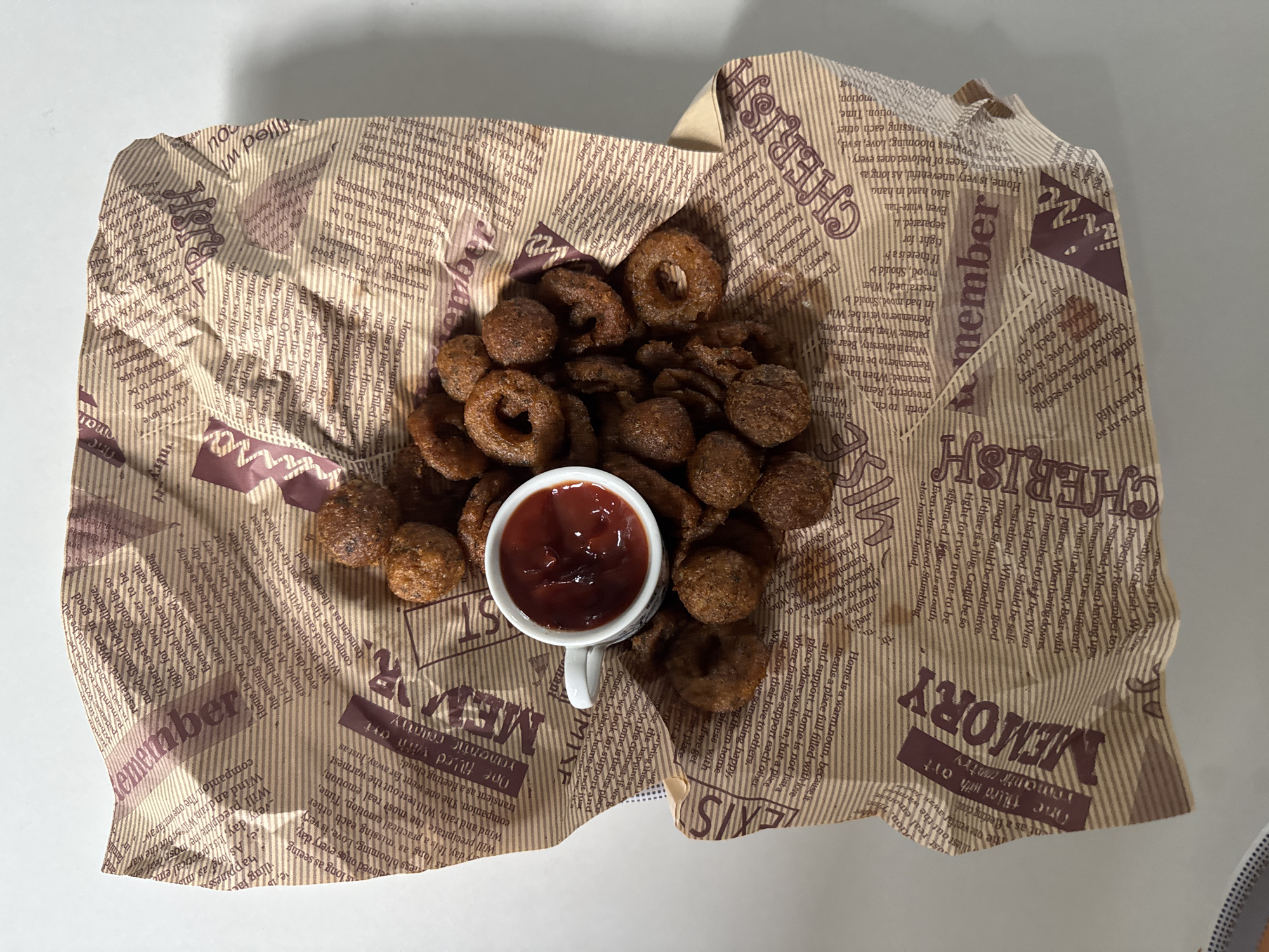 An overhead shot displays a serving of dark brown, fried food items arranged on a crumpled piece of light brown paper, which functions as a liner or wrapper. The food items consist of both ring-shaped and irregularly formed pieces, suggesting a snack like onion rings or a similar fried delicacy. In the center of the food, a small white cup holds a vibrant red sauce, likely ketchup or a dipping sauce.
The paper is extensively printed with text and decorative elements in a darker brown color. Prominent words and phrases visible include "CHERISH," "member," and "MEMORY," often repeated. Other readable fragments convey messages related to home, love, happiness, and endurance, such as "Home is a warm noun, because it means a place full filled with love and support," "When in adversity, Bear with Patience," and "The one filled with romantic country." The text creates a newspaper-like or decorative pattern on the wrapper. The scene is brightly lit, suggesting an indoor setting.