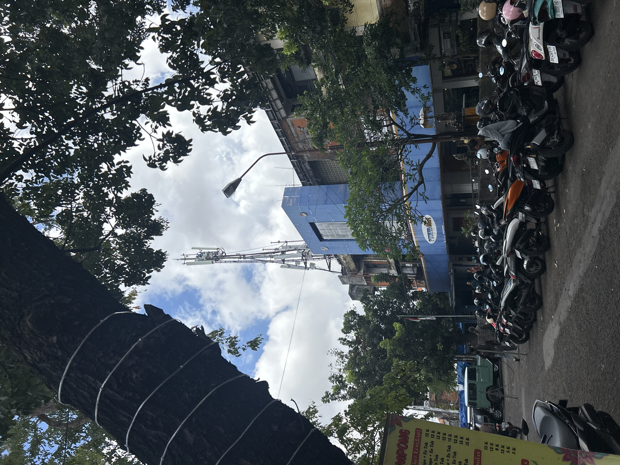The image depicts a street scene in Denpasar, Indonesia, characterized by a row of parked motorcycles, a vintage green Jeep, and a multi-story building with a prominent cell tower. A large tree with exposed roots and a metallic wrap around its trunk dominates the left foreground, its branches extending across the upper portion of the frame. The building, painted blue and yellow, features a cellular transmission tower on its roof, reaching high into the sky. The street is paved, and several individuals are visible, including a man sitting on a motorcycle and others walking or standing near the building. A yellow sign with Indonesian text is partially visible on the left. The sky above is partly cloudy.