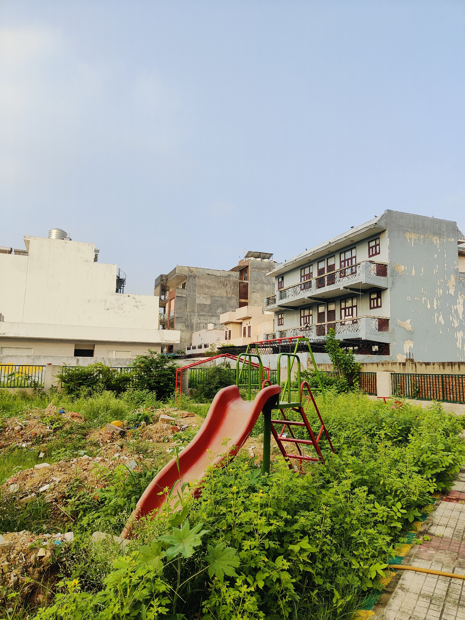 The image captures a neglected children's playground in Gurgaon, India. A red plastic slide is partially visible, set against a backdrop of overgrown green vegetation and piles of construction debris, suggesting a lack of maintenance. In the background, several multi-story residential buildings are prominent. The architecture of these buildings, with their balconies and window styles, is characteristic of urban Indian construction. The sky is clear and light blue, indicating daytime. The scene appears to be a public space that has fallen into disrepair, contrasting with the surrounding residential structures.