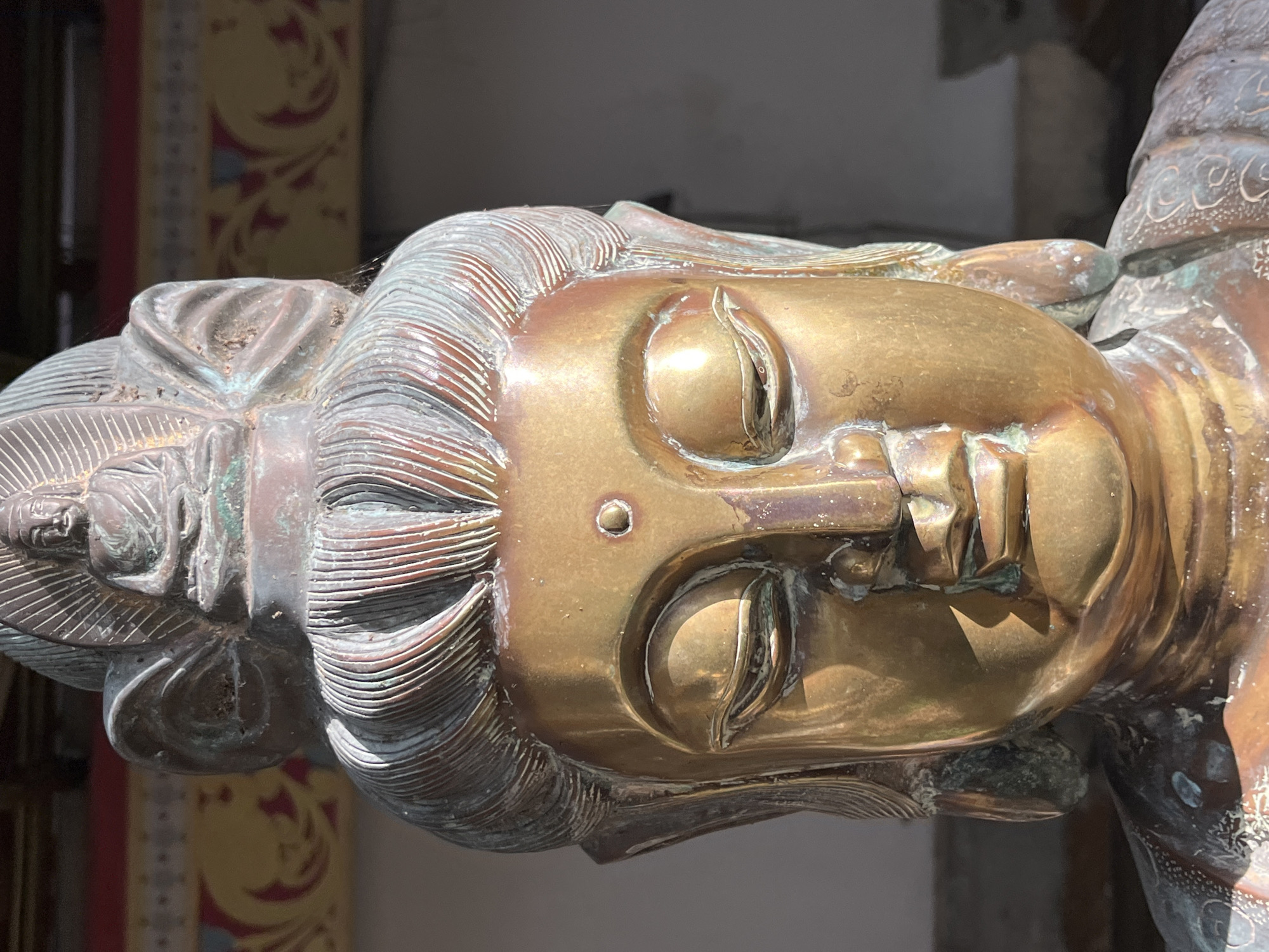 A close-up, eye-level shot shows a weathered bronze statue of a Buddhist deity. The statue's face is golden and illuminated by direct sunlight, highlighting the serene expression with closed eyes, a small urna on the forehead, and pursed lips. The hair is depicted with sculpted waves, and a smaller Buddha figure sits atop the headdress. The bronze shows signs of age with green patina and rough textures on the ears and shoulders. The background is blurred, revealing architectural elements with red and yellow decorative patterns, possibly from a temple or cultural site in Colombo, Sri Lanka.