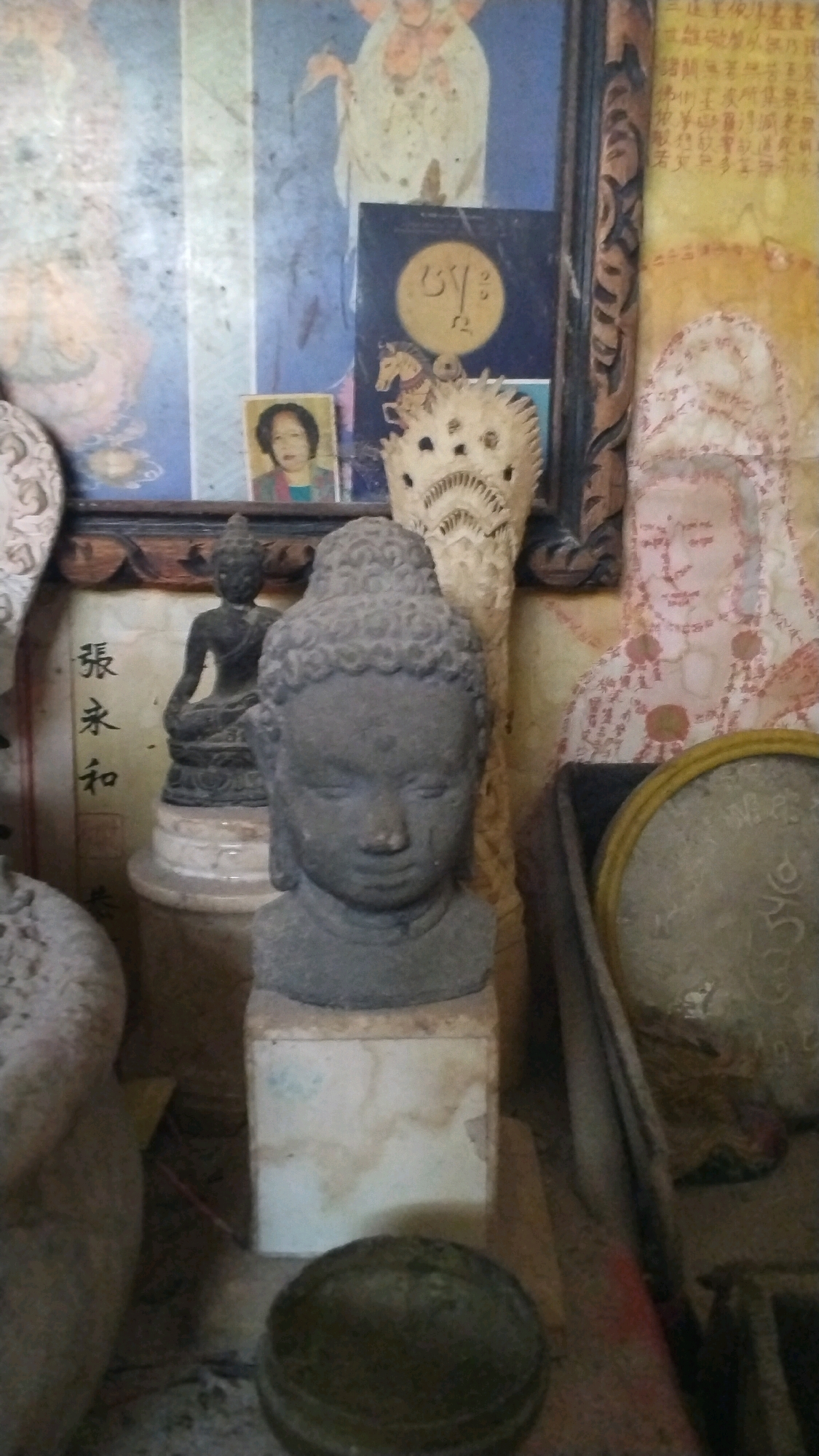 In a dimly lit interior, possibly a shrine or a residential space in Penompo, Indonesia, several devotional and decorative objects are arranged. Dominating the foreground is a weathered stone bust of a Buddha, its serene face tilted slightly downwards. Behind it, a smaller, darker Buddha statue sits in a meditative pose on a pedestal. To the left of the bust, a carved ivory-like object, possibly a decorative finial or mask, is prominently displayed.
Further back, a framed painting depicts a religious figure with outstretched arms, rendered in pale colors with blue and vertical stripes. Below this painting and to the right of the smaller Buddha statue, a small framed photograph shows a woman with dark hair. To the right of the main Buddha bust, a brass-colored circular object, possibly a mirror or decorative plate with calligraphy, rests in a wooden container. The background features a tapestry or painting with intricate patterns and text in an Asian script. The overall scene is cluttered, suggesting a personal collection of religious and artistic artifacts. The lighting is low, casting shadows and highlighting the textures of the stone and carvings.
