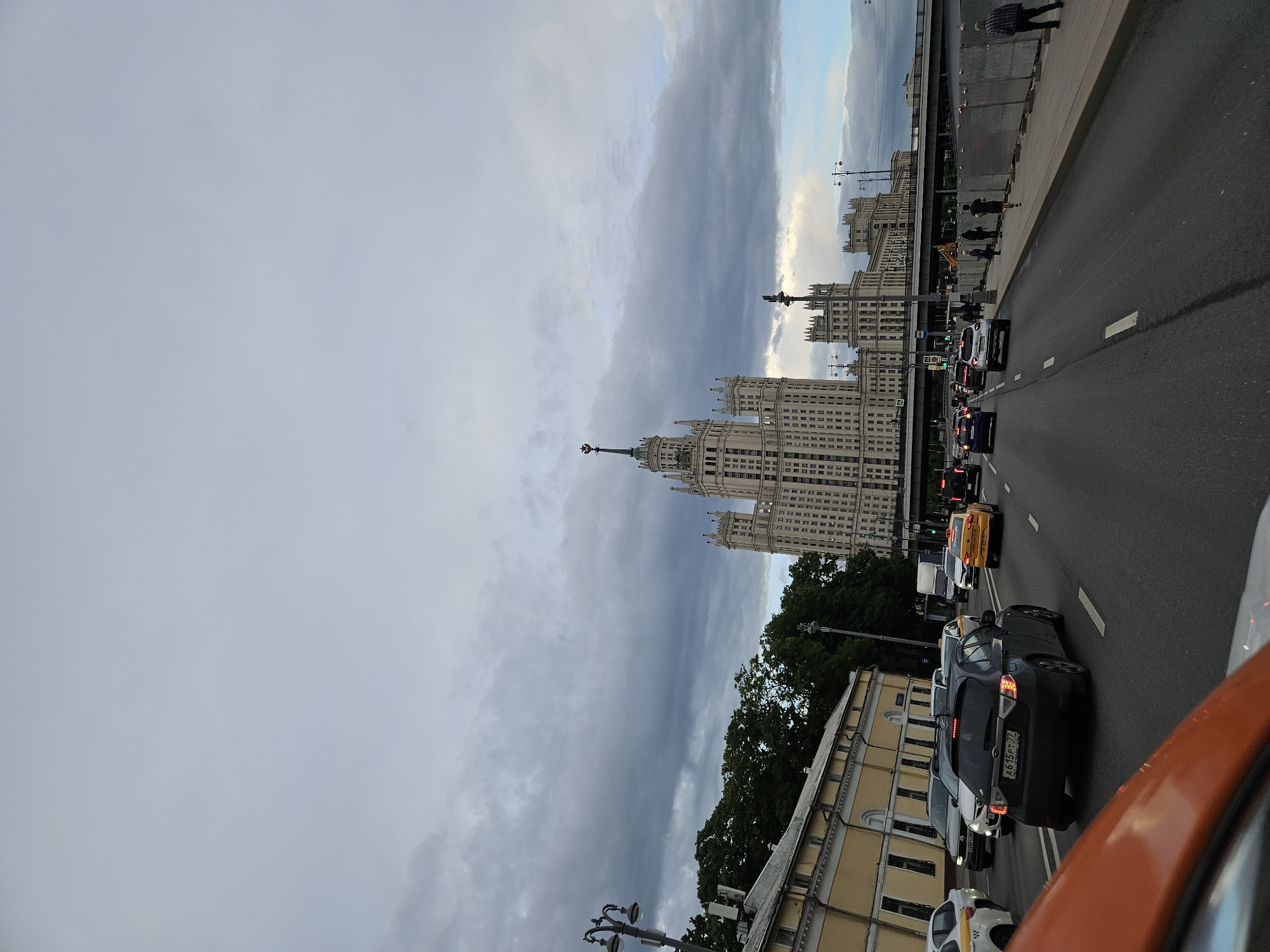 The image captures a street scene in Moscow, Russia, featuring the Kotelnicheskaya Embankment Building, one of the "Seven Sisters" skyscrapers constructed during the Stalinist era. The building dominates the view, rising prominently against a cloudy sky. The street is filled with vehicular traffic, including cars and taxis, suggesting an active urban environment. Pedestrians are visible on the sidewalk adjacent to the road, walking near the bridge. The overall atmosphere appears overcast, with grey tones prevalent in the sky and surrounding cityscape.