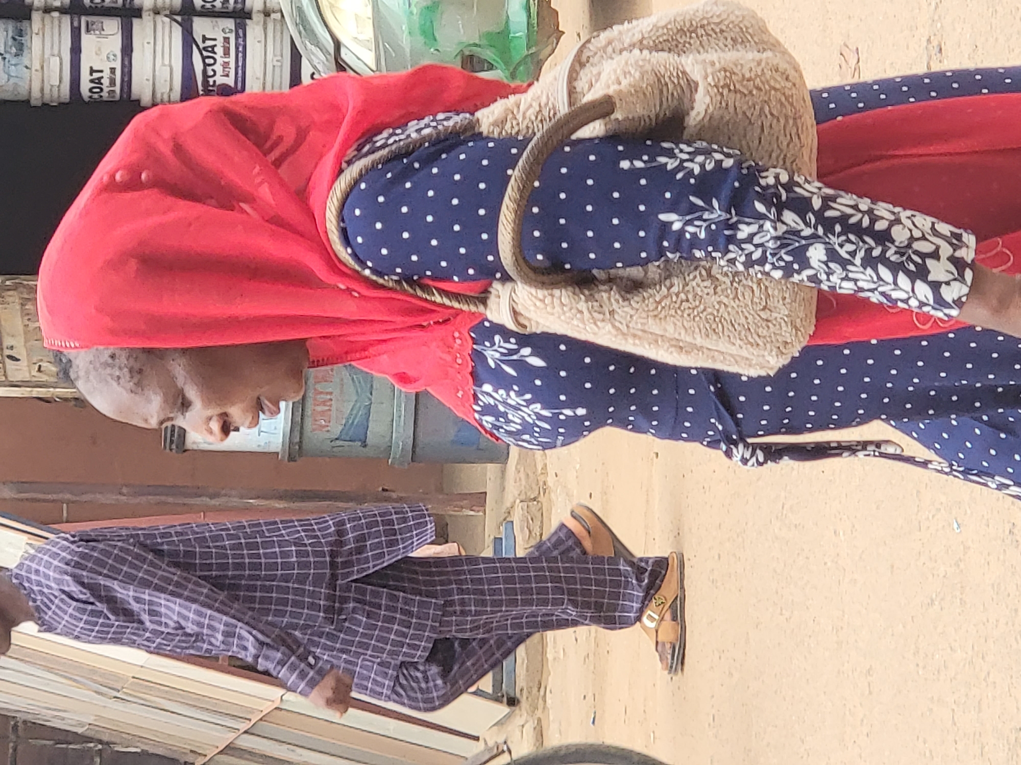 The image captures an outdoor street scene in Maiburiji, Nigeria, featuring two people. A woman, positioned on the right side of the frame, is seen in profile, looking downwards. She wears a bright red head covering, likely a hijab or scarf, and a long blue dress adorned with white polka dots on the main fabric and white floral patterns on the sleeves. A light brown, fluffy shoulder bag with a thick, rope-like strap is slung across her body.
Behind the woman, stacks of cylindrical containers, resembling paint cans, are visible. Text on these containers includes "COAT", "ECOAT", and "Acrylic Emulsion". Green elements, possibly plants or reflections, are also in the background.
On the left, a man is depicted in motion, walking away, though his left profile is partially visible. He is dressed in a matching set of dark blue or purple checkered long-sleeved shirt and trousers. He wears light brown, open-toed sandals, one of which has a small, metallic emblem on its strap. The ground is a light-colored, dusty surface, suggesting an unpaved street or pathway. A partially visible sign behind the man also shows the word "PAINT". The scene is brightly lit, indicating daytime and clear weather, portraying an ordinary street encounter.