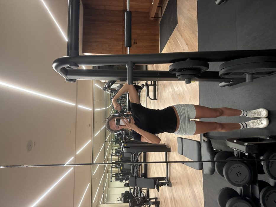 A woman in a black t-shirt and gray shorts poses, flexing her arm in a gym mirror. Her phone is visible in her hand. Dumbbells and weight machines fill the background. The gym has a wood floor and light-colored walls. A weight machine is visible above her.