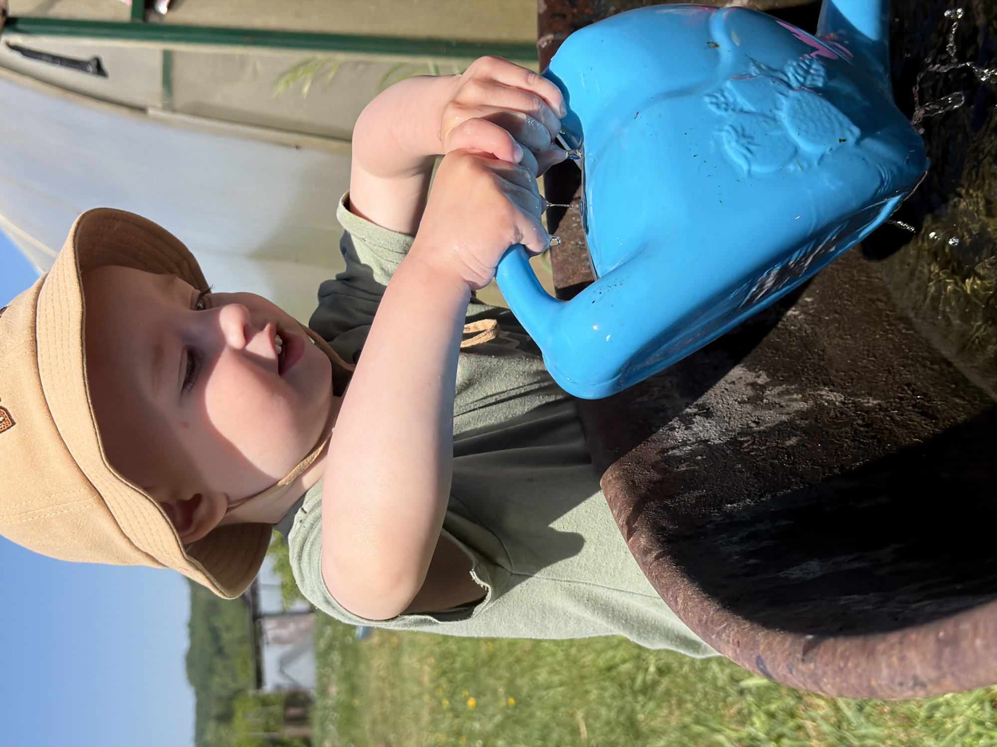A toddler in a tan bucket hat and green shirt is playing with a blue watering can. The child's hands are wet, and water is dripping from the can. The can is positioned over a dark, water-filled basin. The background shows a green field and what appears to be a white greenhouse or tent-like structure. The location is Уральское, Russia.