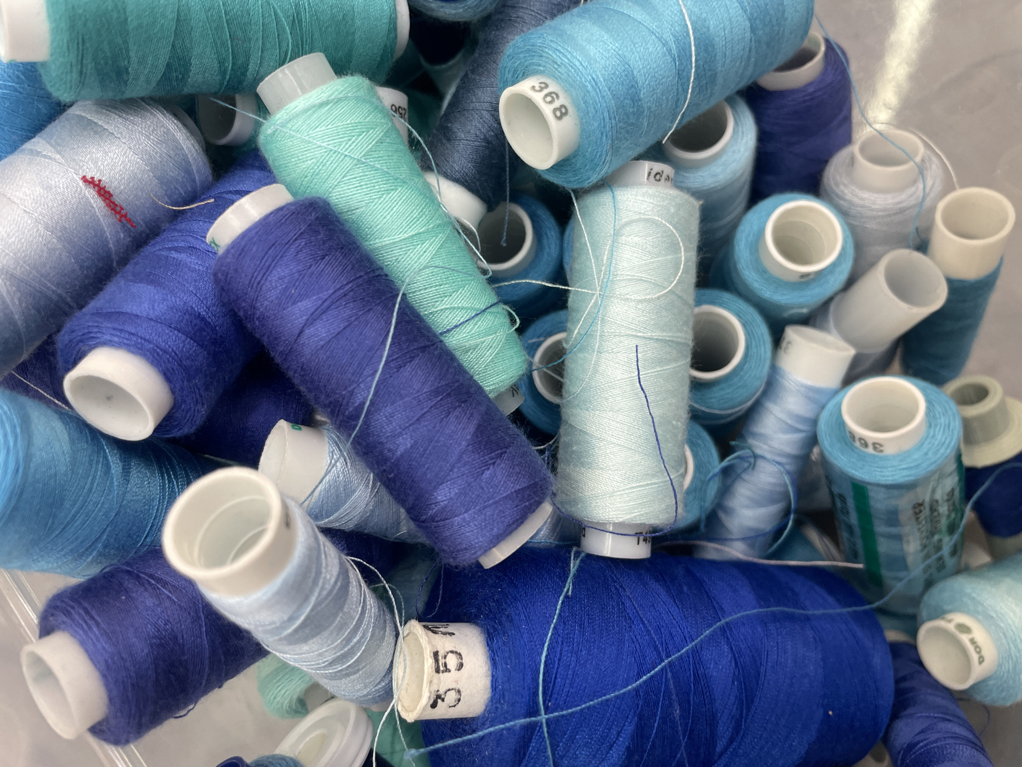 This image is a close-up, high-angle shot of a large collection of spools of thread. The spools are primarily in various shades of blue and teal, with some lighter blues and a few darker blues interspersed. The threads are wound tightly around white plastic spools, and some of the spools have numbers or labels printed on them, such as "351" and "896". The threads themselves are fine and tightly spun. The background is out of focus and appears to be a flat, light-colored surface. The image was taken in Pargolovo, Russia.