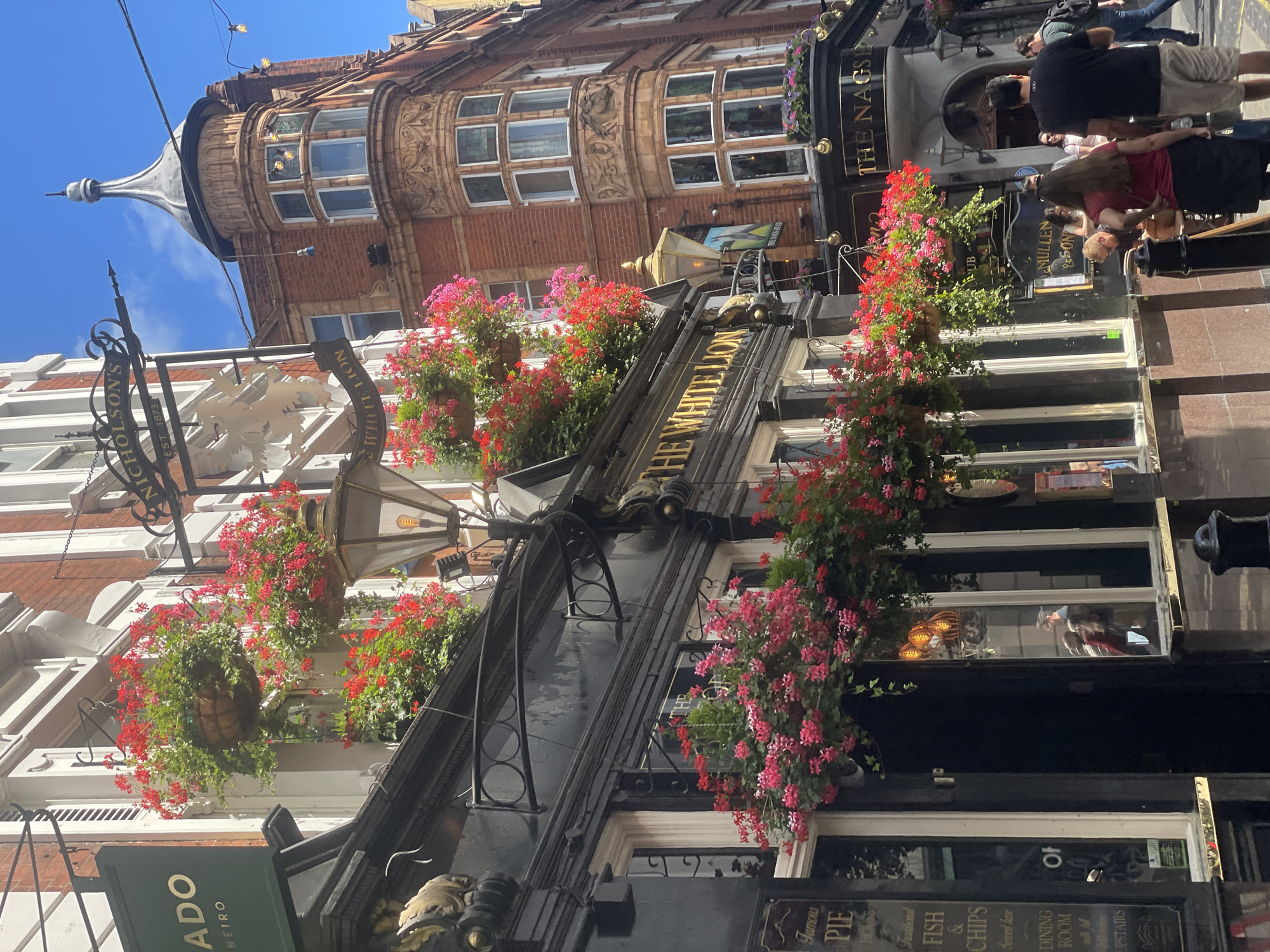 This is a medium shot of a street scene in London, United Kingdom, on a bright, sunny day. The focus is on the architecture of old buildings and a pub. A sign for "NICHOLSON'S" with "EST 1873" is visible, as is a sign for "THE WHITE LION." Another sign reads "THE NAGS HEAD." Hanging baskets overflowing with red and pink flowers adorn the facade of the buildings, creating a vibrant contrast with the brick and stone architecture. People are walking in the background, some appearing to enter or exit the pub. The overall impression is a charming and historic streetscape, characteristic of a bustling London area. Visible text includes pub names and establishment details, along with a sign for "Traditional FISH & CHIPS" and "DINING ROOM."