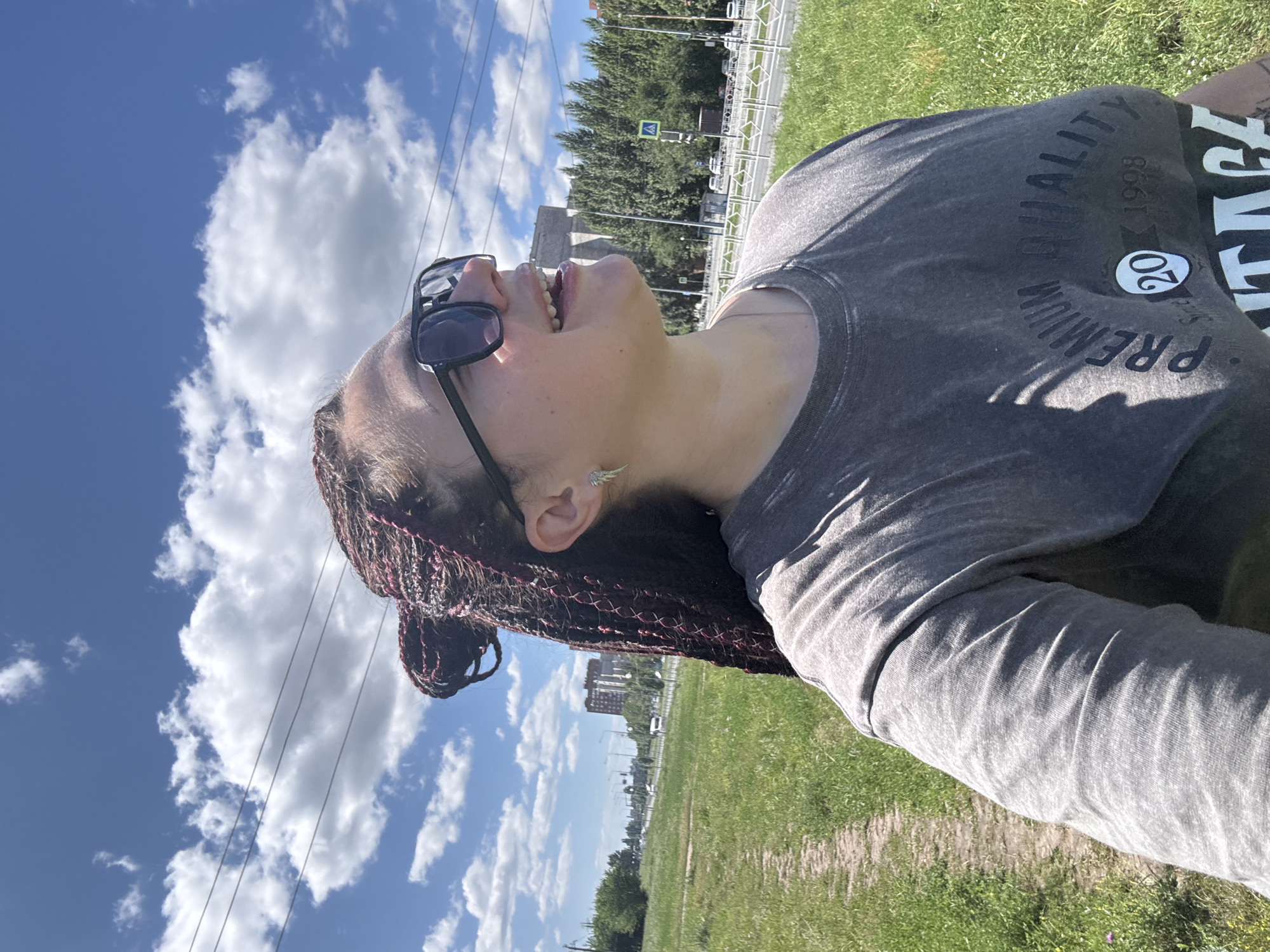 A medium shot shows a woman with dark braided hair and sunglasses looking up and to the right, smiling. She is wearing a grey t-shirt with "PREMIUM QUALITY 1998" printed on it. She is standing on a grassy area next to a dirt path, with a road, trees, and buildings in the background. The sky is blue with white clouds. This scene is likely located in the city of Tolyatti, Russia, given the presence of urban infrastructure and typical vegetation for the region.