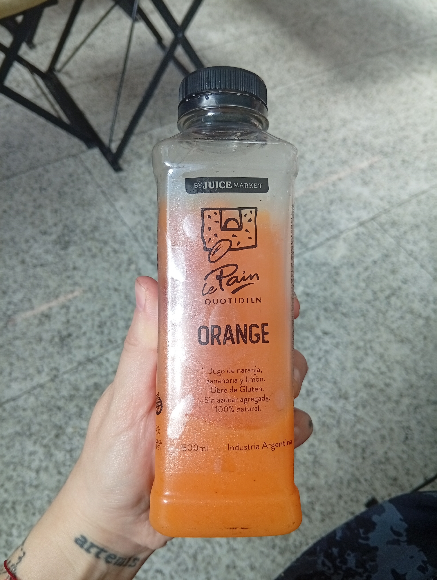A person is holding a bottle of orange juice, labeled "Le Pain Quotidien," with the description in Spanish: "Jugo de naranja, zanahoria y limón" (orange, carrot, and lemon juice). The bottle also states "Industria Argentina," and "500ml." The person has a tattoo on their wrist that reads "artemis." The background appears to be a tiled floor with a black metal chair. Based on the label and the Argentine indication, the location is potentially Aeropuerto Internacional Ezeiza, Argentina.