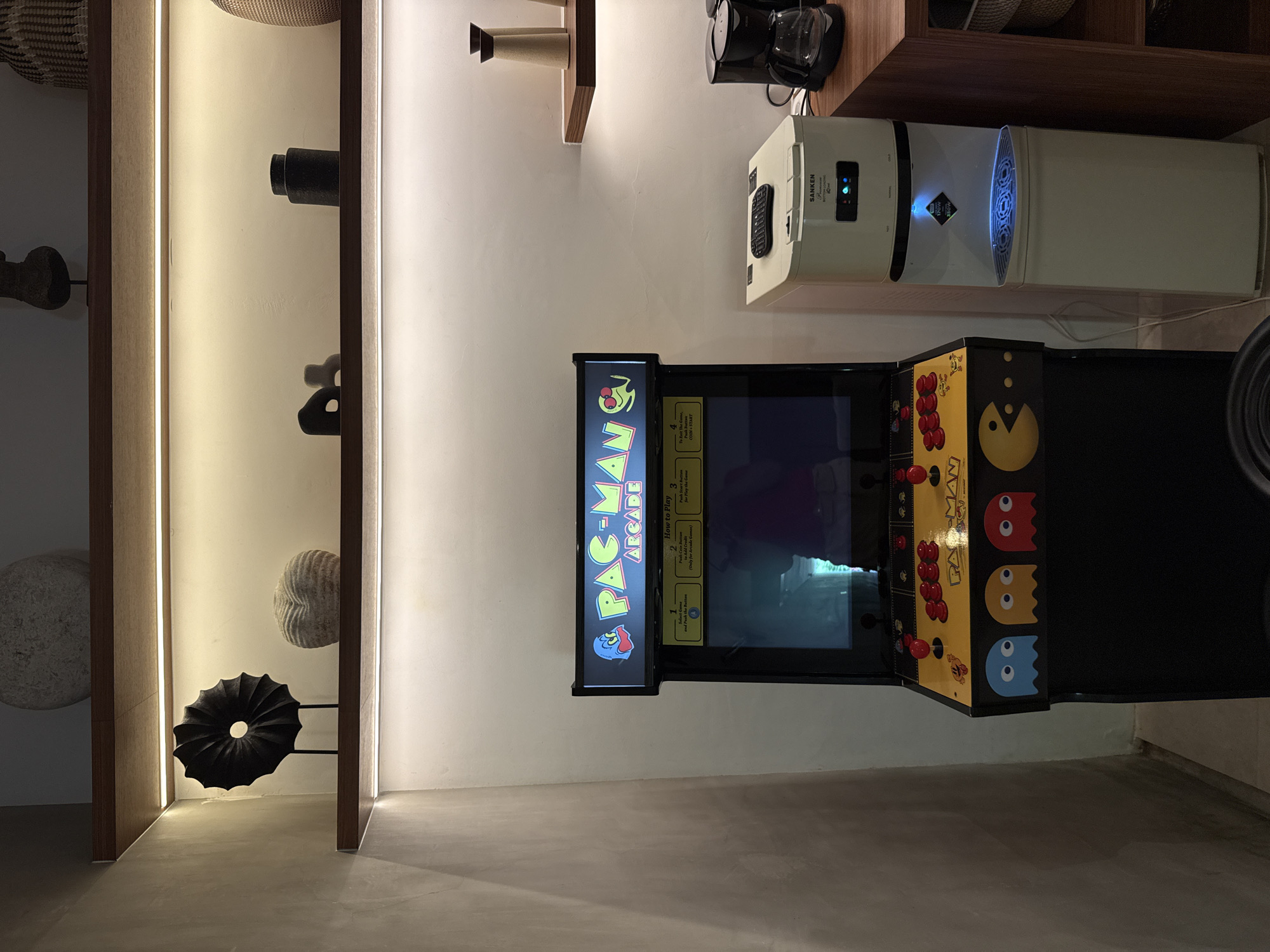 Here's a matter-of-fact description of the image:
The image presents an interior scene in a vertical format. A Pac-Man arcade game is the focal point, displaying a retro aesthetic with its classic graphics and button controls. To the left of the arcade game is a shelving unit that is built into the wall, featuring various decorative objects. Above the arcade game on the wall is an appliance that could be a water dispenser or filter. The lighting in the room is a combination of natural and artificial, with illuminated strips highlighting the edges of the shelving unit. The image depicts a location in the city of Ubud District, Indonesia.