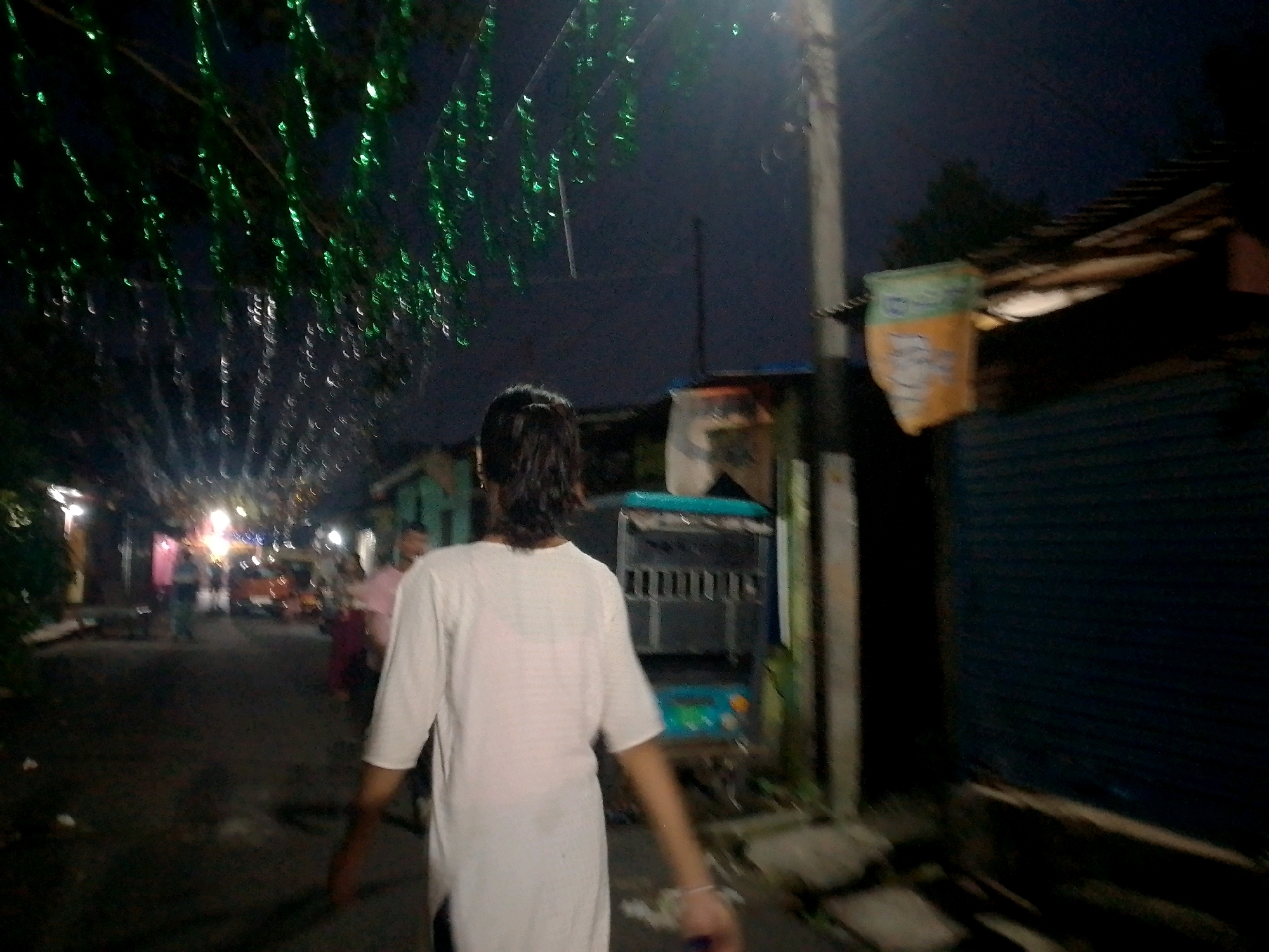 The image depicts a dimly lit street scene at night in Siliguri, India. A person, viewed from behind, is walking away from the camera. They are wearing a white t-shirt. The street is lined with buildings and appears to be a commercial or residential area. Green festive lights hang overhead, casting a glow on the scene. Other individuals are visible in the background, some appearing to be walking or standing. There is a vehicle, possibly a cart or small truck, on the right side of the street. The overall atmosphere is that of a local street during an evening or night time event.