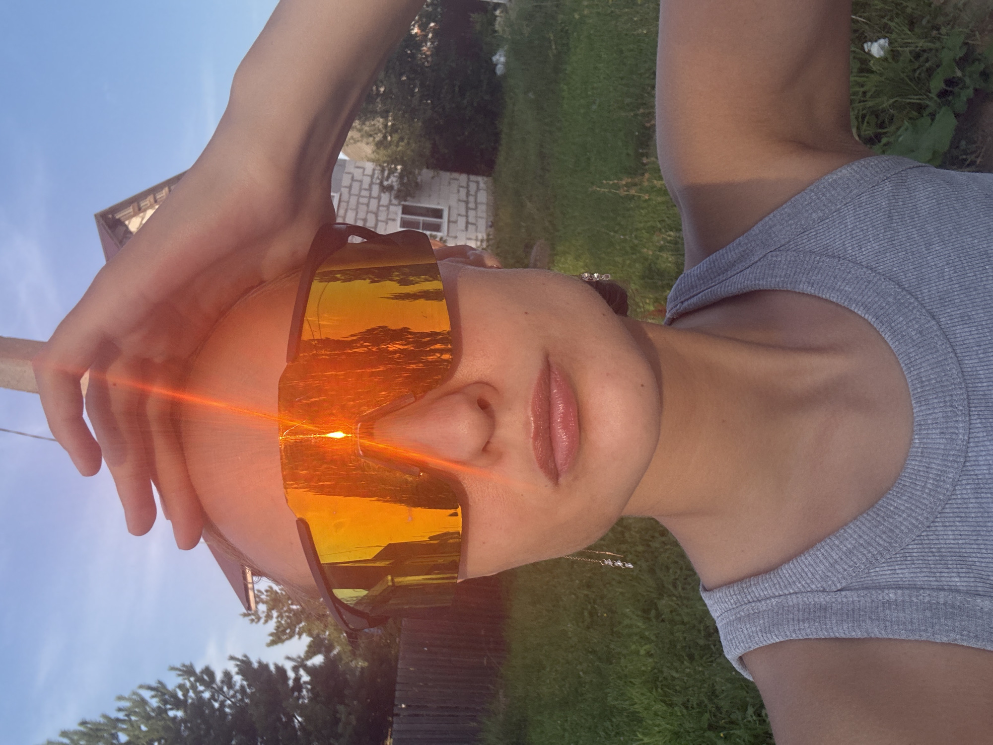 The image is a selfie of a person wearing large, orange-tinted sunglasses. The person's face is mostly visible, with their lips and nose in focus. Their hand is raised and resting on their head. The sunglasses reflect the setting sun, creating a bright orange glow that also illuminates the person's forehead. In the background, a grassy area with trees and a small building can be seen. The sky is clear and blue. The image appears to be taken outdoors during sunset, likely in a rural or suburban area given the background. The overall mood seems relaxed and summery. There is no visible text in the image. The location context is given as Podshivalovo, Russia.