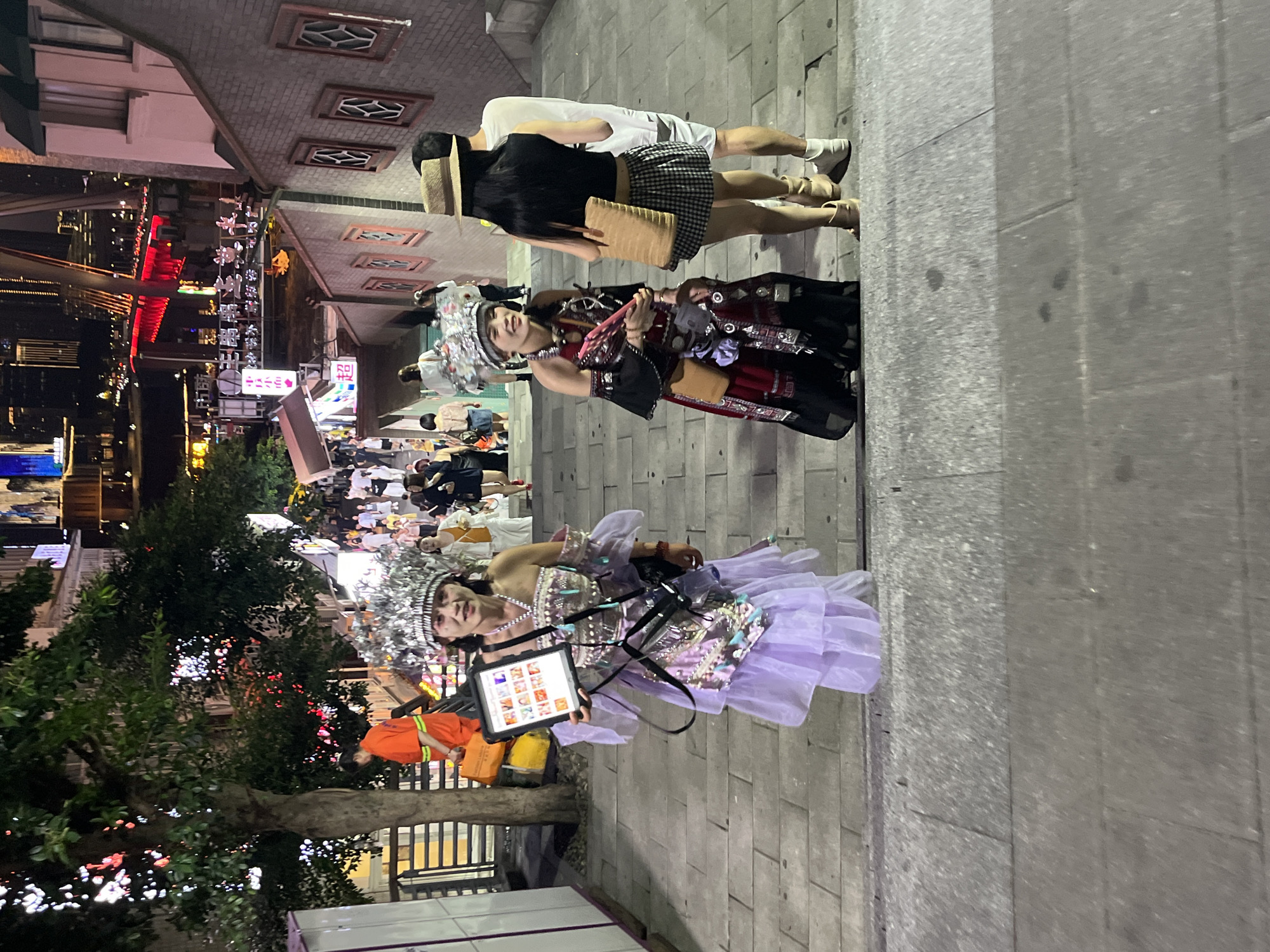 This is a medium-shot, eye-level, outdoor, nighttime, vertical photograph of two women dressed in ornate traditional costumes, standing on a wide, grey stone staircase. They are in the Yuzhong District, China, likely a tourist area, as indicated by the background cityscape with illuminated buildings and bridges, and a crowd of pedestrians.
The woman on the left is wearing a shimmering silver headdress adorned with intricate patterns and dangling elements. Her dress is a layered, voluminous gown with a pale lavender, sheer outer skirt over a more solid, decorative underlayer. She holds a tablet computer in her left hand, displaying a grid of what appear to be small images. A dark shoulder bag is slung across her body, and another bag is attached to her waist. Her right hand is resting on the tablet.
The woman on the right is also wearing a decorative headdress, though it appears slightly different from the first woman's. Her dress is a dark, long-sleeved garment with red and black patterned fabric and intricate embellishments. A tan-colored purse is slung across her chest, and a small tan bag is attached to her waist. She is looking to her left, and her right hand is partially obscured by her clothing.
Behind the two women, another couple is visible. The woman in front is wearing a straw hat and a black tank top with a black and white checkered skirt, holding a tan tote bag. The man behind her is wearing a white t-shirt and dark shorts, with white sneakers. Further up the stairs, more people are walking and standing, some wearing casual clothing, others in what seem to be other traditional outfits or performance attire.
The staircase itself is constructed of large, grey stone slabs. The background shows a dynamic, urban environment at night, with tall buildings, vibrant lights, and a bridge spanning a waterway. The overall atmosphere suggests a lively night market or cultural event.