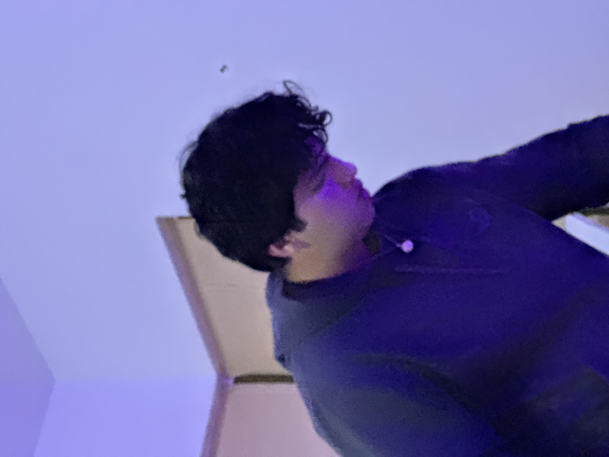 A low-angle, blurry image shows a person with dark, curly hair and tan skin. They wear a dark blue or purple jacket or sweatshirt. A small white object, potentially an earbud, is visible near their collar. The background consists of a light purple or blue wall and a portion of a light-colored doorway or corner. The location is not discernible as Oaxaca City, Mexico from the visual details provided.