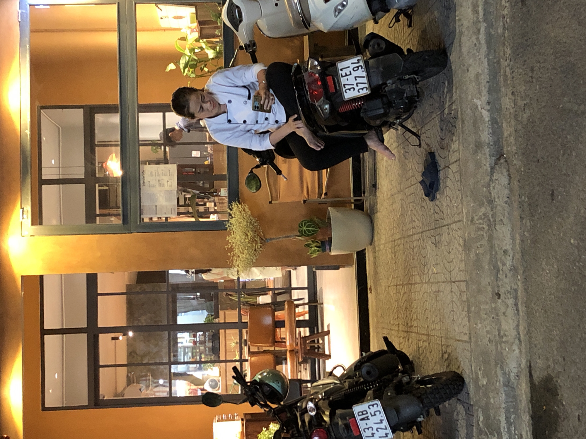 A woman in a white chef's coat is sitting on a black scooter parked on a tiled sidewalk in front of a building with large windows and an orange facade. She is looking down at her phone, which she holds in her right hand. Her left hand is resting on the scooter's seat. The scooter has a license plate that reads "37-E1 377.94". A pair of dark blue flip-flops are on the sidewalk to the left of the scooter. In the background, through the windows, there appears to be a cafe or restaurant interior with tables and chairs. A smaller motorcycle is parked to the left of the woman and scooter. The lighting suggests it is evening or nighttime, with warm light illuminating the scene from the building and streetlights. This scene is characteristic of a street scene in Đà Nẵng, Vietnam, where scooters are a common mode of transportation and outdoor seating is prevalent.