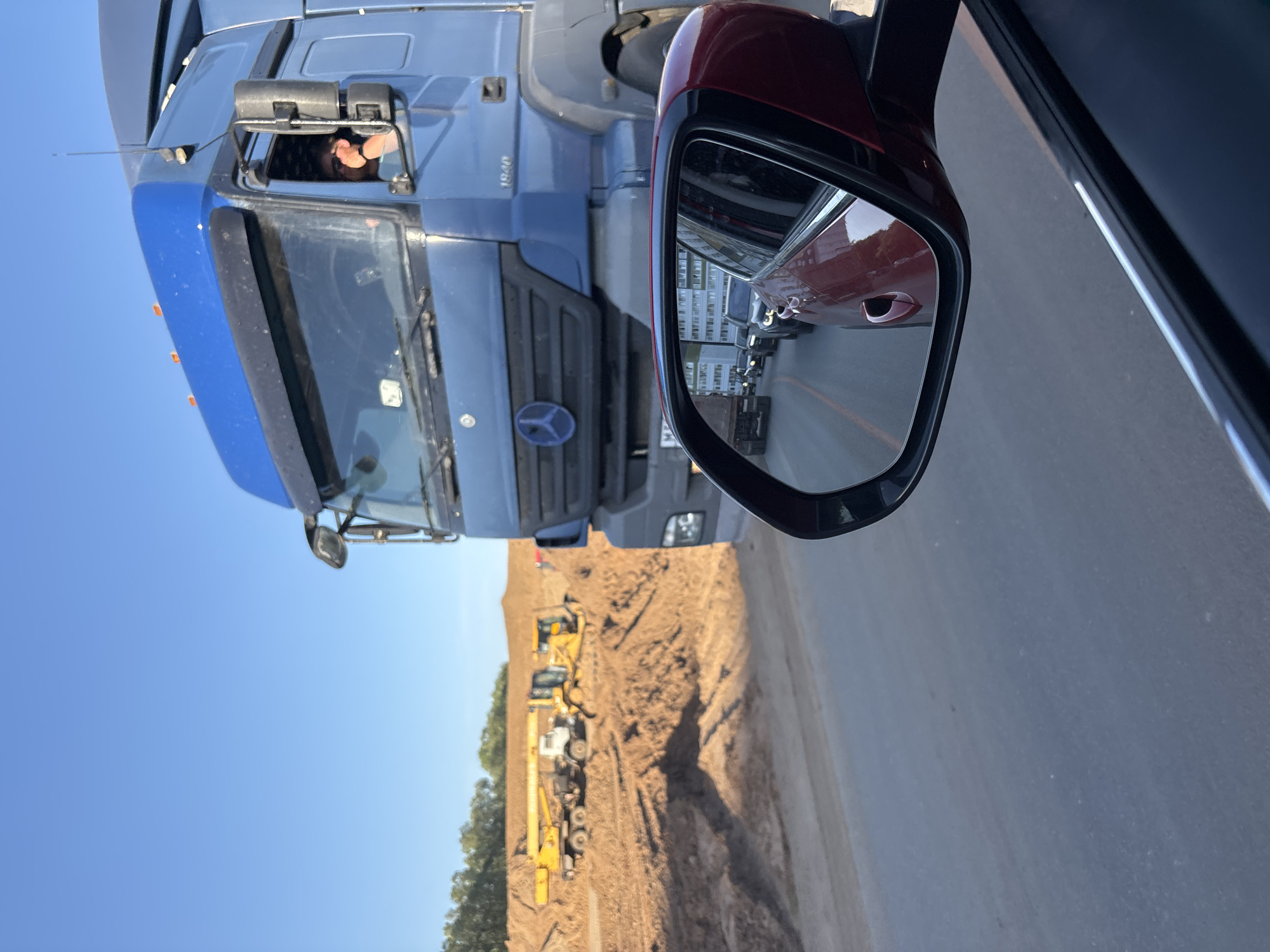A roadside view shows a large blue Mercedes-Benz truck with the driver visible in the cab. The truck is beside a passenger vehicle, where a red car side mirror can be seen. Reflected in the mirror are other cars and buildings, suggesting traffic behind the photographer's vehicle. The terrain to the side of the road appears to be an active construction or excavation site, with large yellow machinery like excavators and cranes present. The environment includes a clear, blue sky above the construction zone.
I am not certain about the location in the city of Kabanikha, Russia, as it cannot be determined from the visual information provided.