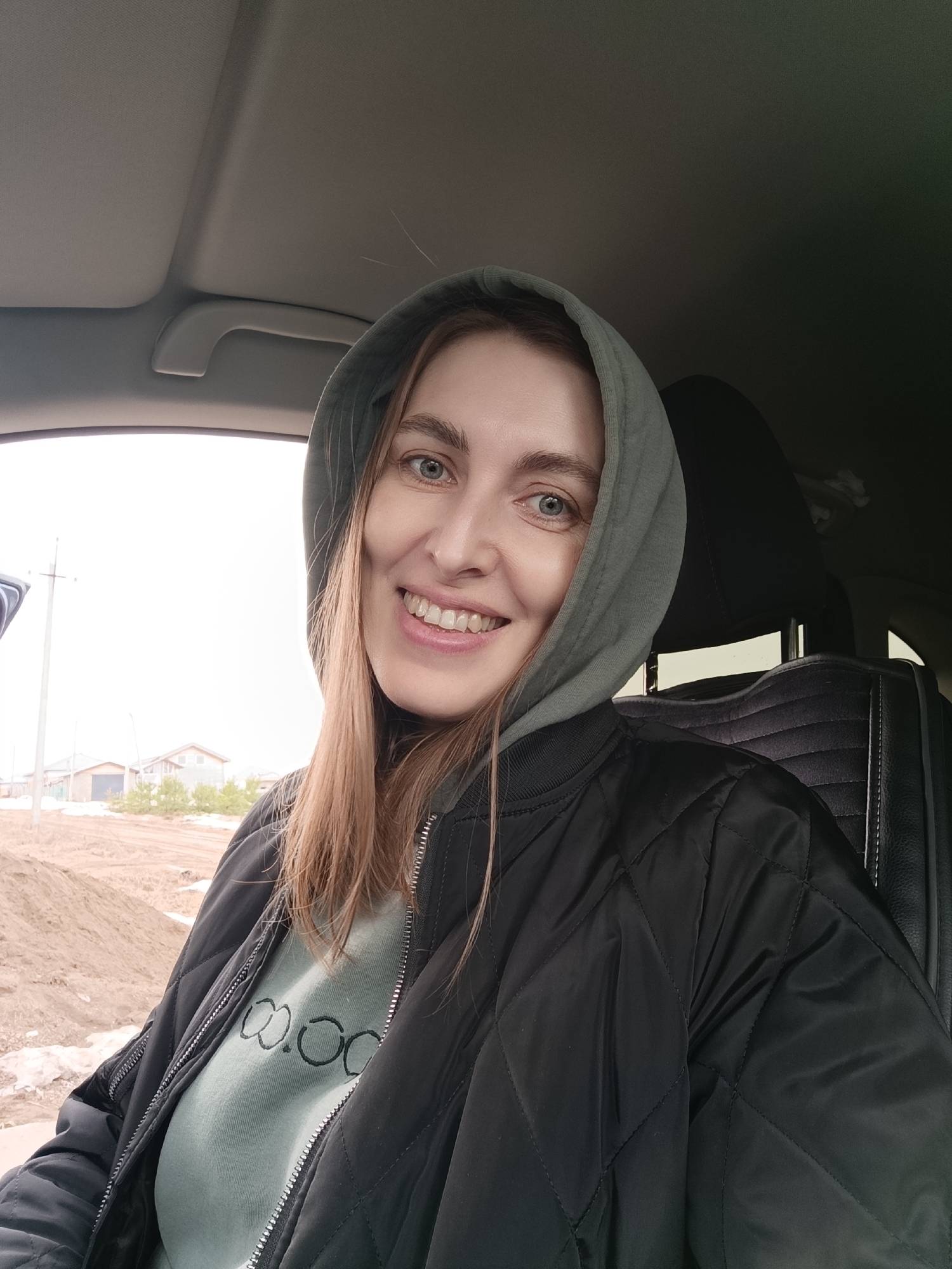 A woman is sitting in a car, smiling at the camera. She has light brown hair, blue eyes, and is wearing a light green hoodie and a black quilted jacket. The car's interior is visible, including the dashboard and seats. Outside the car window, a partially constructed area with houses and bare ground is seen. It appears to be Chaikovsky, Russia.
