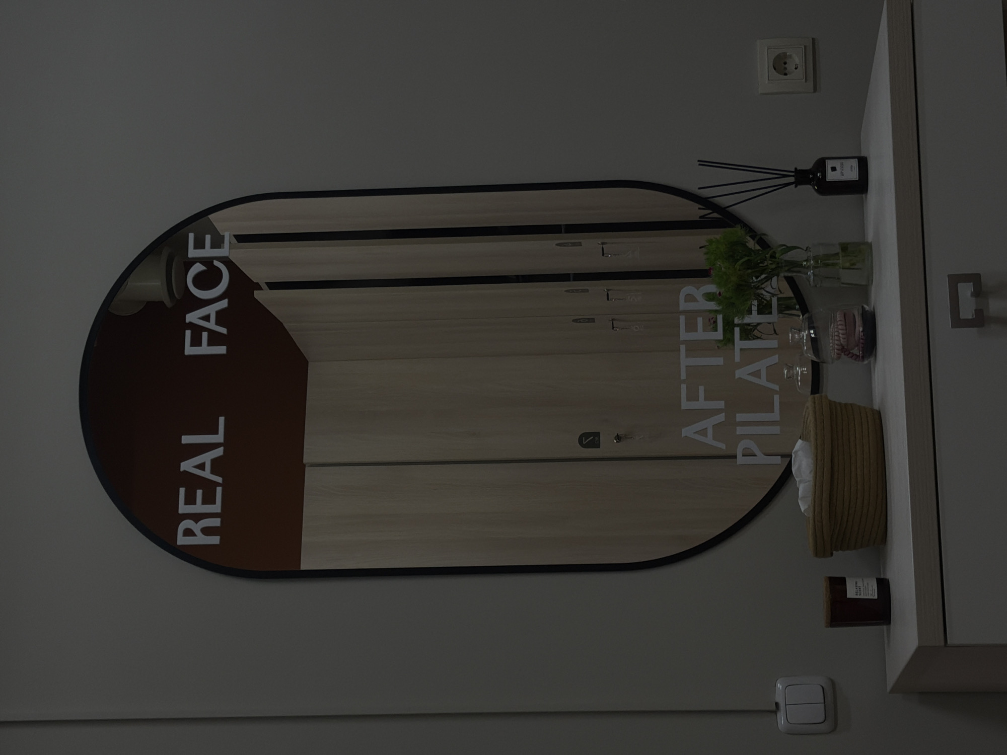 The image shows a wall-mounted oval mirror in a room, with the words "REAL FACE" and "AFTER PILATES" vertically arranged on opposite sides of the mirror's surface. The mirror reflects the interior of a closet or cabinet. To the right of the mirror is a shelf or countertop on which various items are placed, including a reed diffuser, glass vases, a small woven basket with white fabric inside, and a candle. A light switch is visible beneath the shelf, close to the bottom of the image. The wall is painted a neutral gray color, with an electrical outlet above the shelf. The overall setting is a modern, minimalist interior space. There is no information available about the location in Yerevan, Armenia.