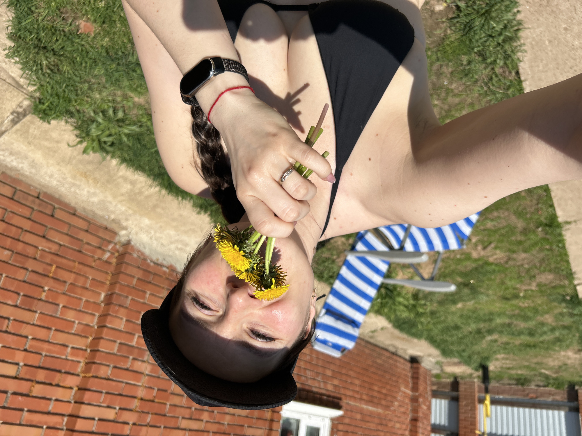 A woman in a black bikini lies on her back outdoors, holding a bouquet of dandelions to her nose. She wears a black cap and a smartwatch on her left wrist. A red bracelet is also visible on her left wrist. A blue and white striped beach chair is nearby. The background includes a brick wall, grass, and a portion of a building. The location is unspecified and there is no evidence of Rusalevka, Russia.