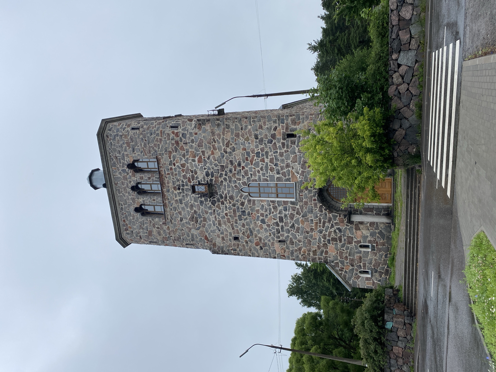 The image shows the Church of the Transfiguration in Priozersk, Russia. The church is a tall, stone tower with a square base and a octagonal top. The stone is of various colors, including gray, brown, and red. The church has several windows, some of which are arched and others are rectangular. The church is surrounded by trees and a stone wall. In the foreground, there is a crosswalk on a wet asphalt road. The sky is overcast. There are no people visible in the image.