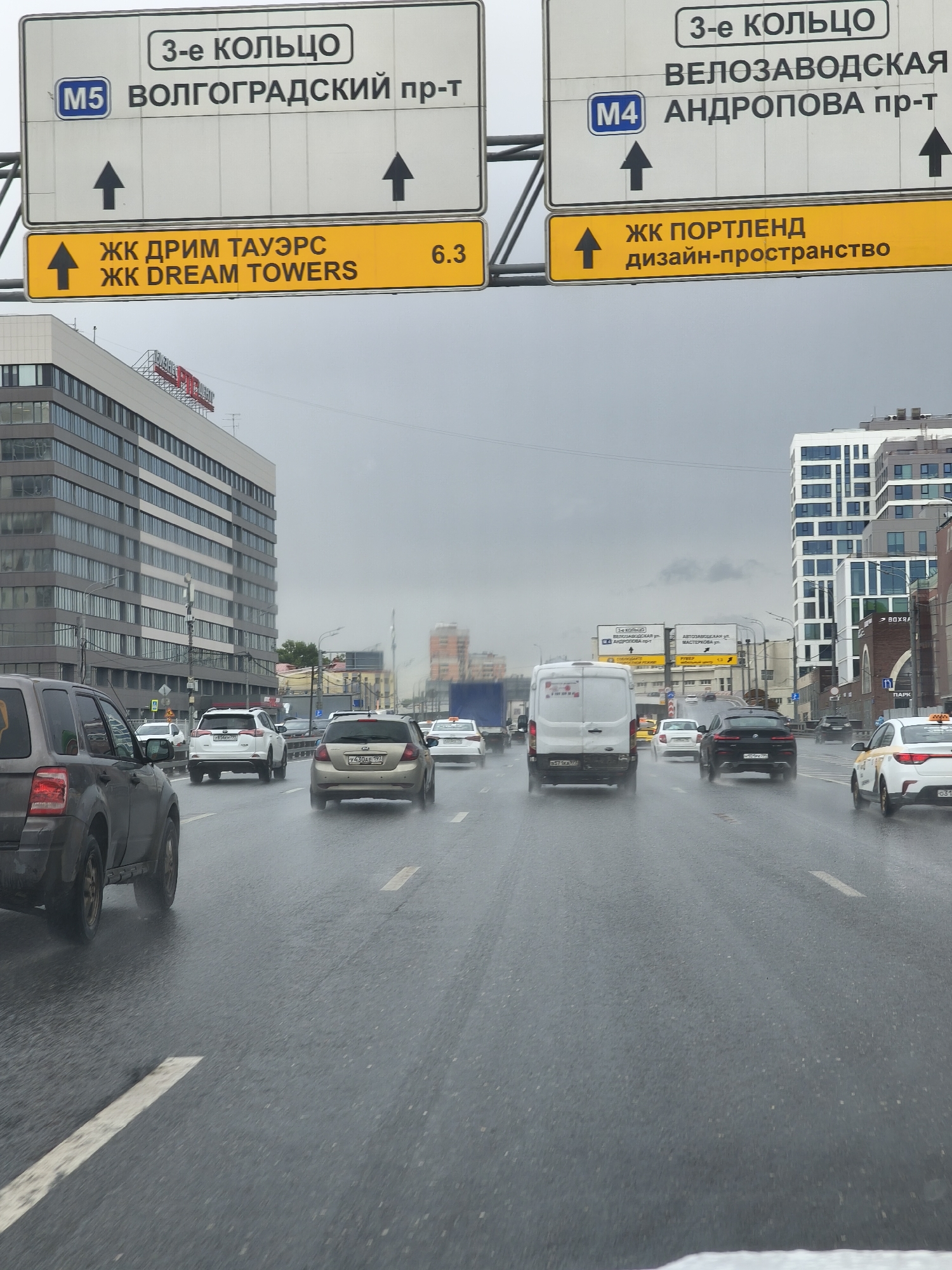 The image depicts a street scene in Moscow, Russia, taken on a wet or rainy day. The road is asphalt and shows signs of being wet from recent or ongoing rain. Multiple lanes of traffic are visible, occupied by various vehicles including SUVs, sedans, vans, and a taxi. Overhead road signs indicate the direction to the "3rd Ring Road," specifically pointing towards Volgogradsky Prospekt (M5) and Andropova Prospekt (M4), near the Velozavodskaya area. Another sign indicates the "Dream Towers" residential complex is 6.3 kilometers in the indicated direction. The architecture in the background includes a mix of modern and older buildings. The sky is overcast.