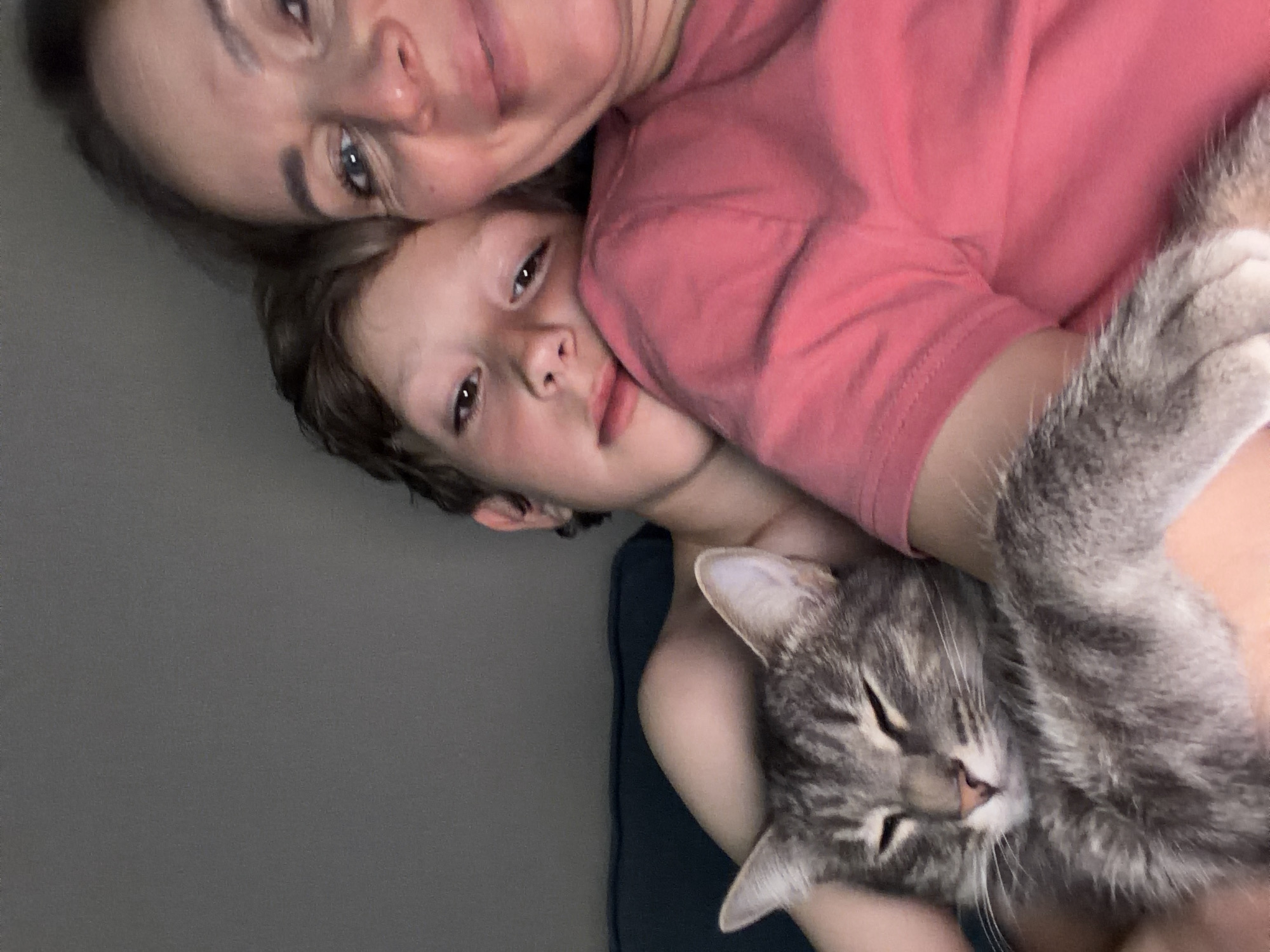 A woman, a boy, and a cat are lying together. The woman and boy are looking at the camera. The cat is nestled against the boy's body, with its eyes closed. The woman is wearing a pink shirt. The background appears to be a grey wall. The location is Chaikovsky, Russia.