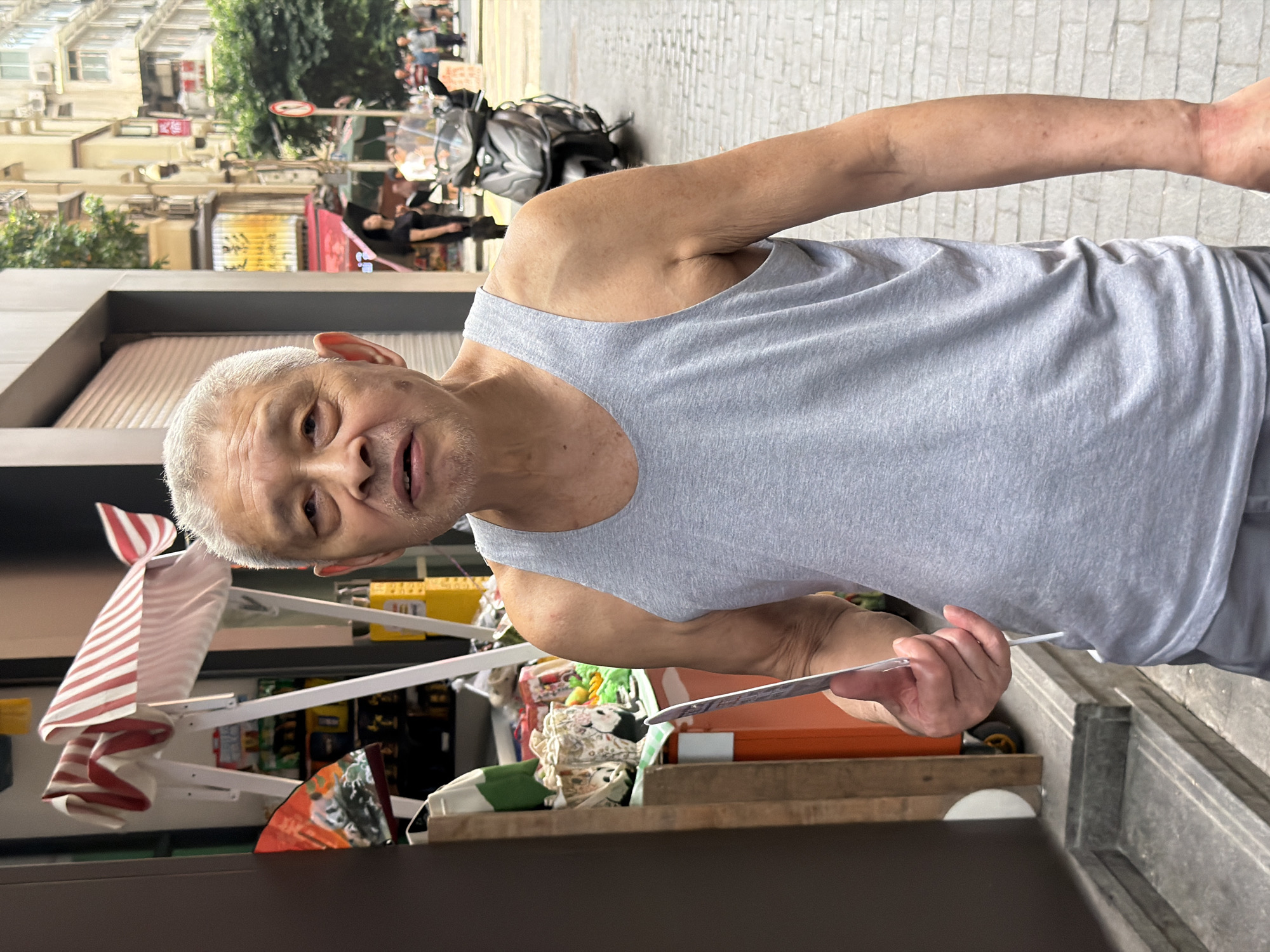 An elderly East Asian man with short, gray hair and light facial stubble stands outdoors in a narrow street. He is wearing a gray tank top and gray shorts. He is holding a flat, white object in his right hand, with his thumb and index finger gripping its edge. To his left, a storefront displays a striped awning and various goods. Behind him, a motorcycle is parked on the brick-paved street, and people are walking in the background. The buildings in the background are multi-story structures. The scene appears to be set in Yuzhong District, China.