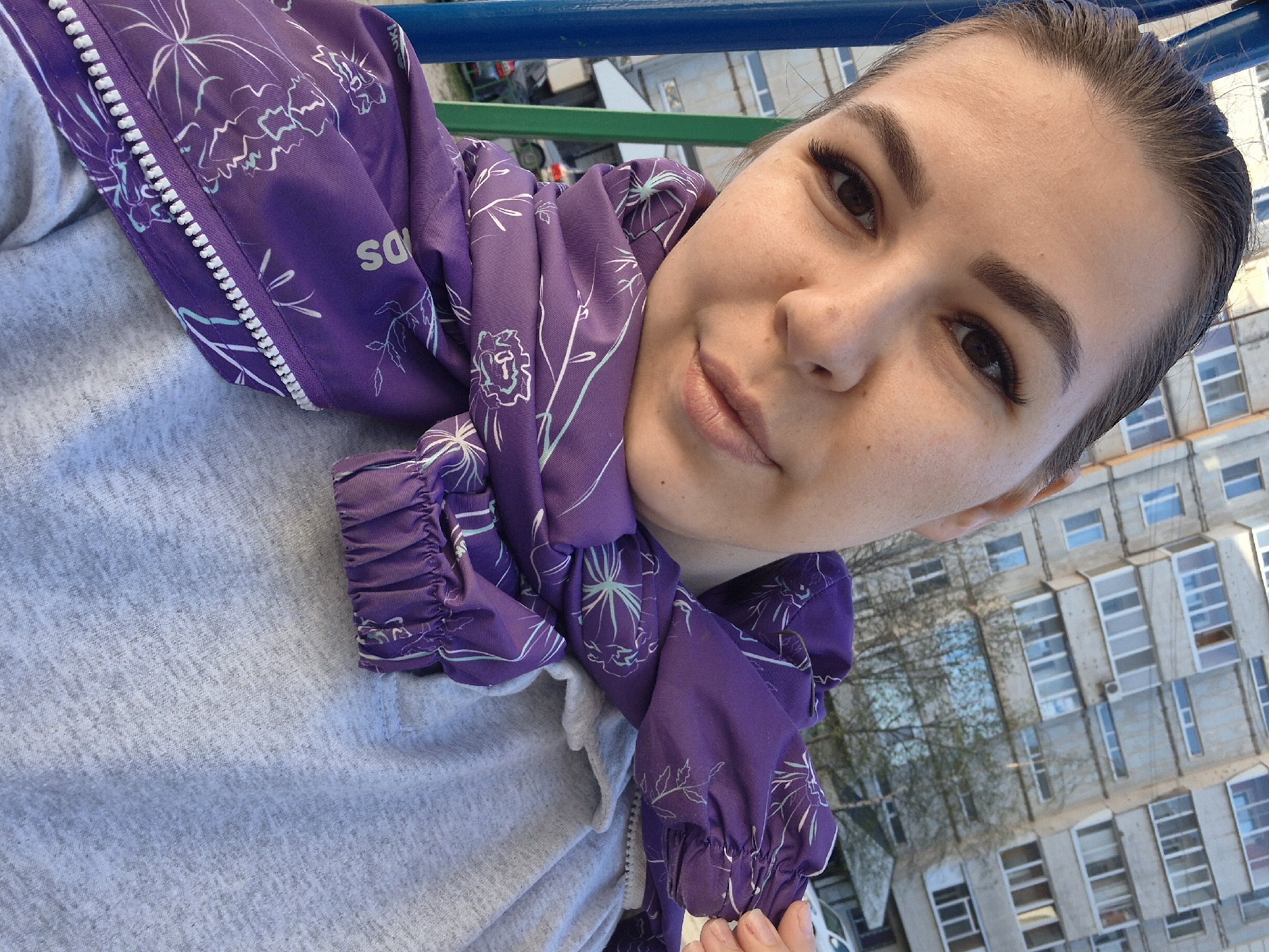 A close-up, canted selfie depicts a young woman in an urban environment. She is fair-skinned with brown eyes, and her hair is slicked back. The woman is wearing a light gray sweatshirt and a purple jacket patterned with white floral designs, casually draped around her neck. The jacket's zipper is visible.
The backdrop features a multi-story building with a repetitive pattern of windows. Bare tree branches are visible in front of the building. The scene appears to be outdoors, with a glimpse of a blue and green structure overhead. The city of Tobolsk, Russia, is not discernable from the available visual information.