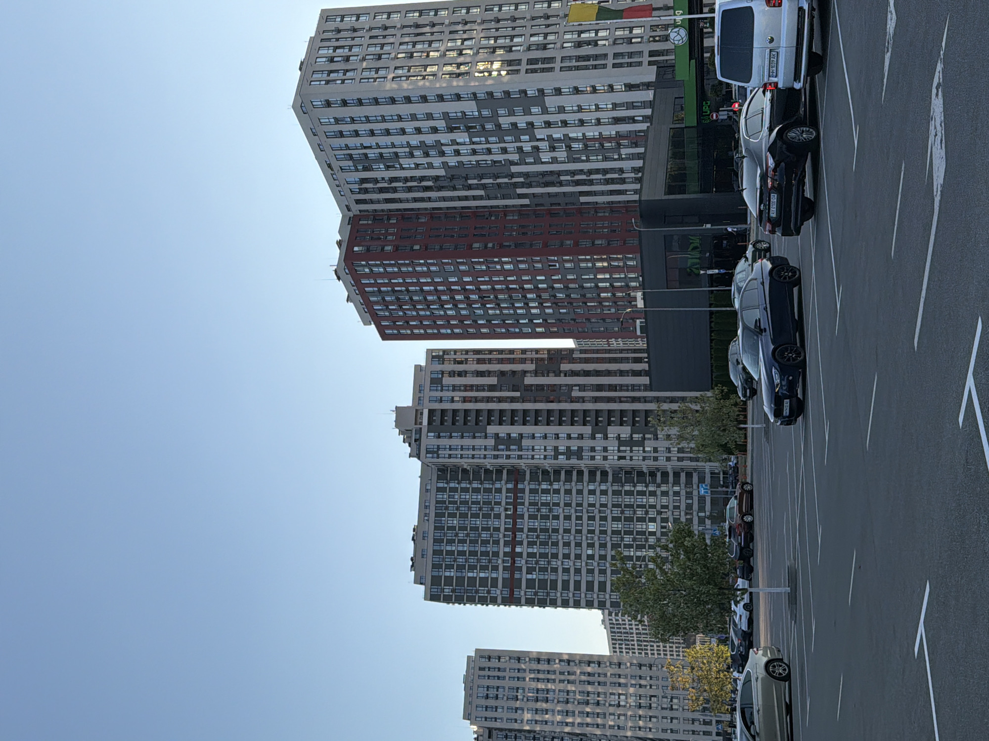 This is a low-angle, full-shot photograph of a parking lot in front of several large, modern apartment buildings. The sky is clear and blue, with the sun casting a warm glow on the buildings. The buildings are a mix of light gray, dark gray, and red, with numerous windows. In the foreground, the asphalt parking lot is marked with white lines. Several cars are parked in the lot, including a silver sedan on the left, a dark blue sedan and a black sedan in the middle, and a white van on the right. A few trees are visible in the middle ground. The scene appears to be in Kyiv, Ukraine, likely a residential area with commercial spaces on the ground floor of the buildings.