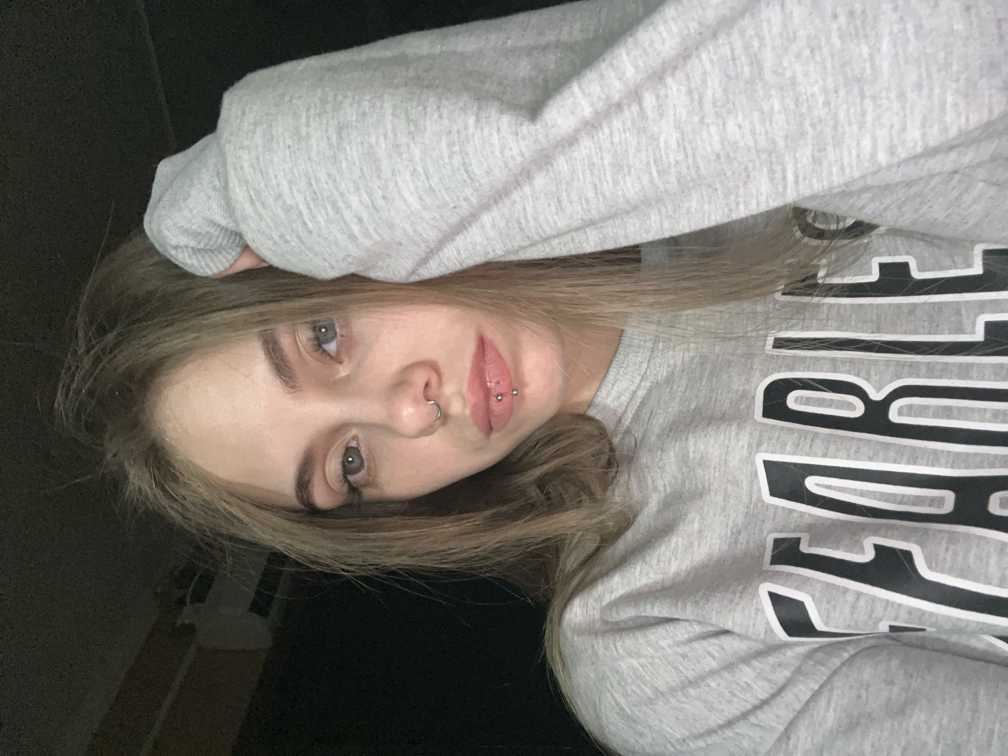 A young woman with light brown hair and blue eyes is posing for a selfie. She is wearing a grey sweatshirt with black and white lettering, which is partially visible. She has piercings in her nose and lower lip. Her left hand is resting on her head. The background is dark and indistinct. The location is unknown.