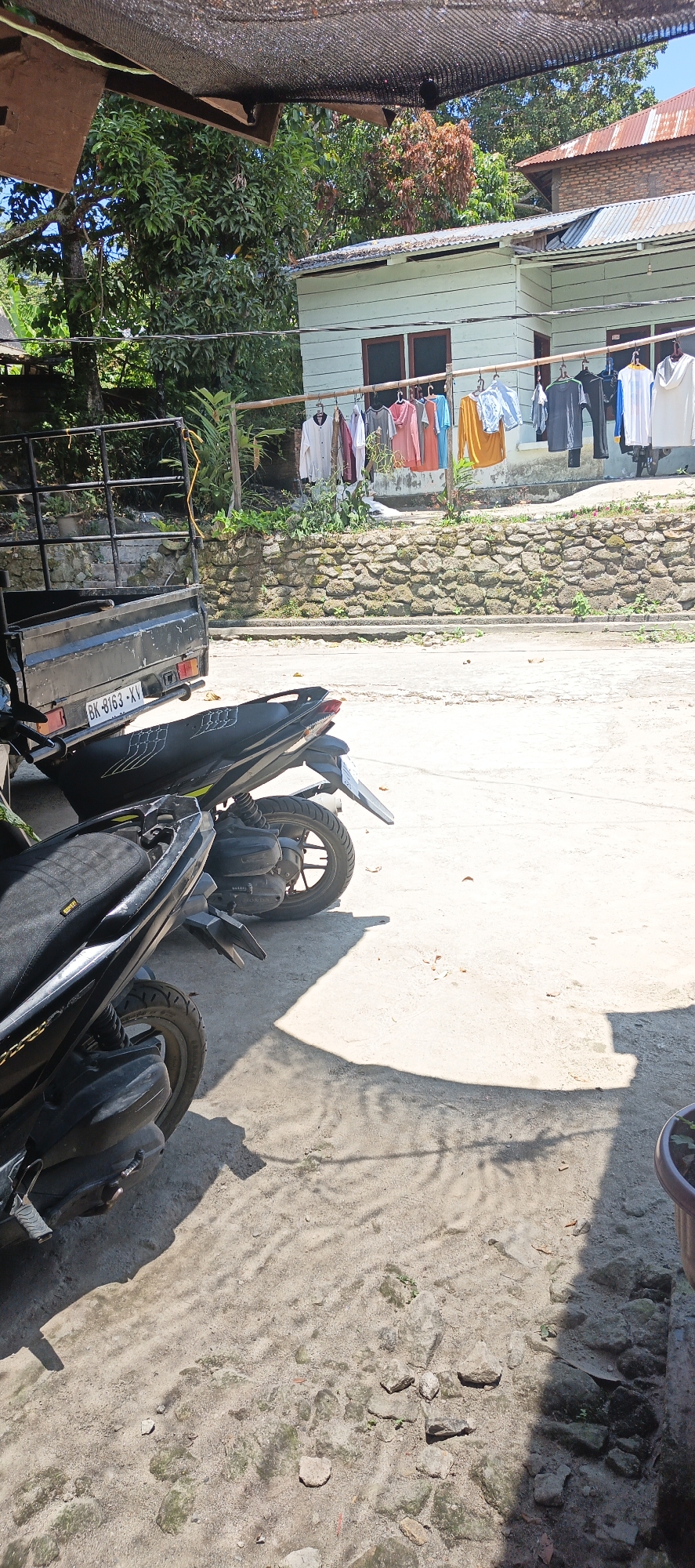 In Cinta Dame, Indonesia, a scene unfolds under a thatched roof structure, likely a carport or outdoor shelter. In the foreground, two black motorcycles are parked side-by-side. The rear motorcycle, a Yamaha Mio, is partially visible, displaying its license plate which reads "BK 8163 XV". The front motorcycle is more prominently featured, its black seat with a yellow "FLEX" sticker facing the viewer. To the left, a black flatbed truck is parked, its side facing the motorcycles. Behind the vehicles, a stone wall lines a slightly elevated area. Beyond the wall, a light-colored house with wooden siding and two dark-paned windows is visible. Clothes are hung on a clothesline in front of the house, including a white long-sleeved shirt, several t-shirts in various colors (pink, orange, yellow, blue, black), and a pair of dark pants. The background is filled with lush green trees and a glimpse of a rusted tin roof on another building. The ground is a mix of concrete and dirt, with scattered leaves and small stones. The overall impression is a residential area in a tropical climate, with everyday life activities such as laundry and parking of vehicles.