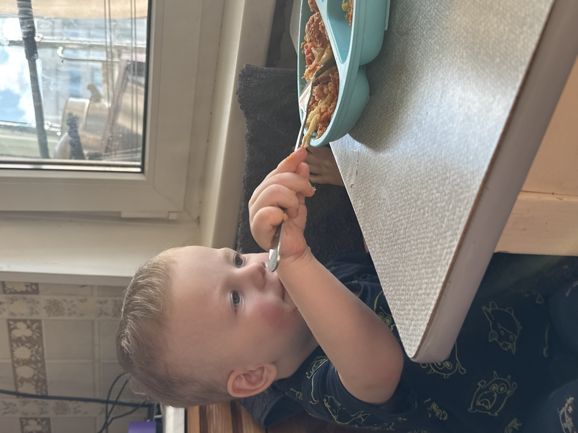 A young child sits at a light-colored table, eating from a turquoise plate with a fork. The child has fair hair and rosy cheeks, wearing a dark blue shirt with light-colored cartoon figures. A window is visible in the background, showing a cloudy sky and buildings, characteristic of the architecture of Saint Petersburg, Russia. A tile wall is visible behind and to the left of the child.