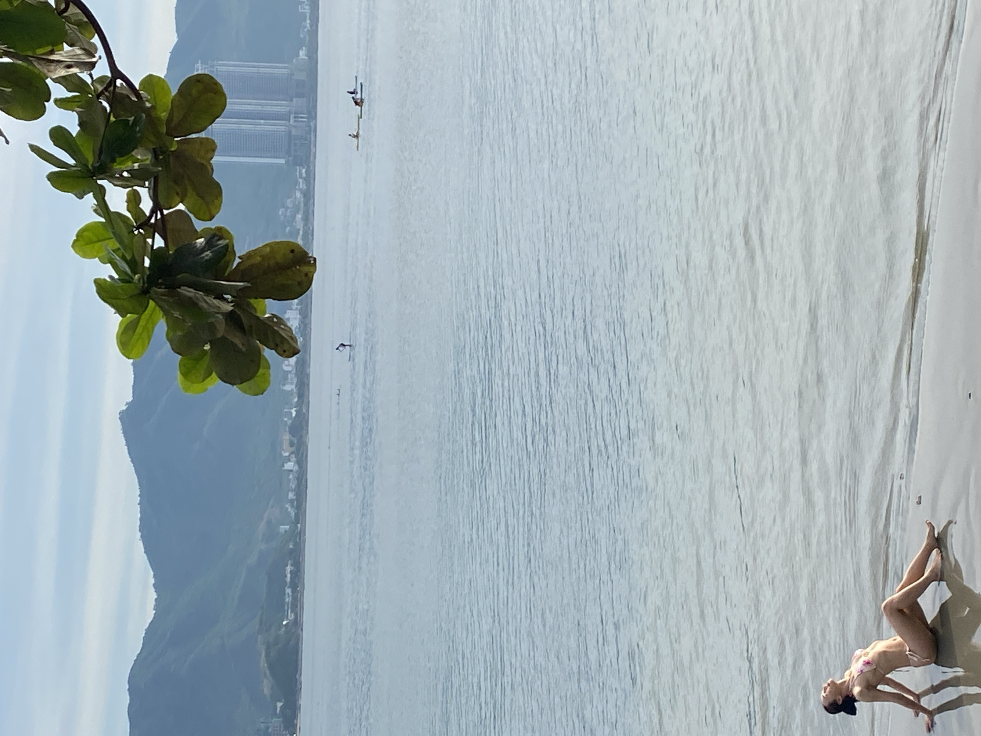 A woman in a bikini sits on a sandy beach with gentle waves lapping at the shore, facing towards the water and leaning back with her head tilted up towards the sky. Her arms are resting on the sand behind her, supporting her upper body. In the background, the calm ocean stretches out towards a distant coastline with lush green mountains. A few individuals are visible in the water, some on paddleboards and others in kayaks, enjoying recreational activities. The sky above is a pale blue with a few wispy clouds. A branch with green leaves hangs from the top right corner of the frame, partially obscuring the view of the distant city buildings and mountains, which suggest the location is Nha Trang, Vietnam. The overall atmosphere is serene and peaceful, depicting a tranquil day at the beach.