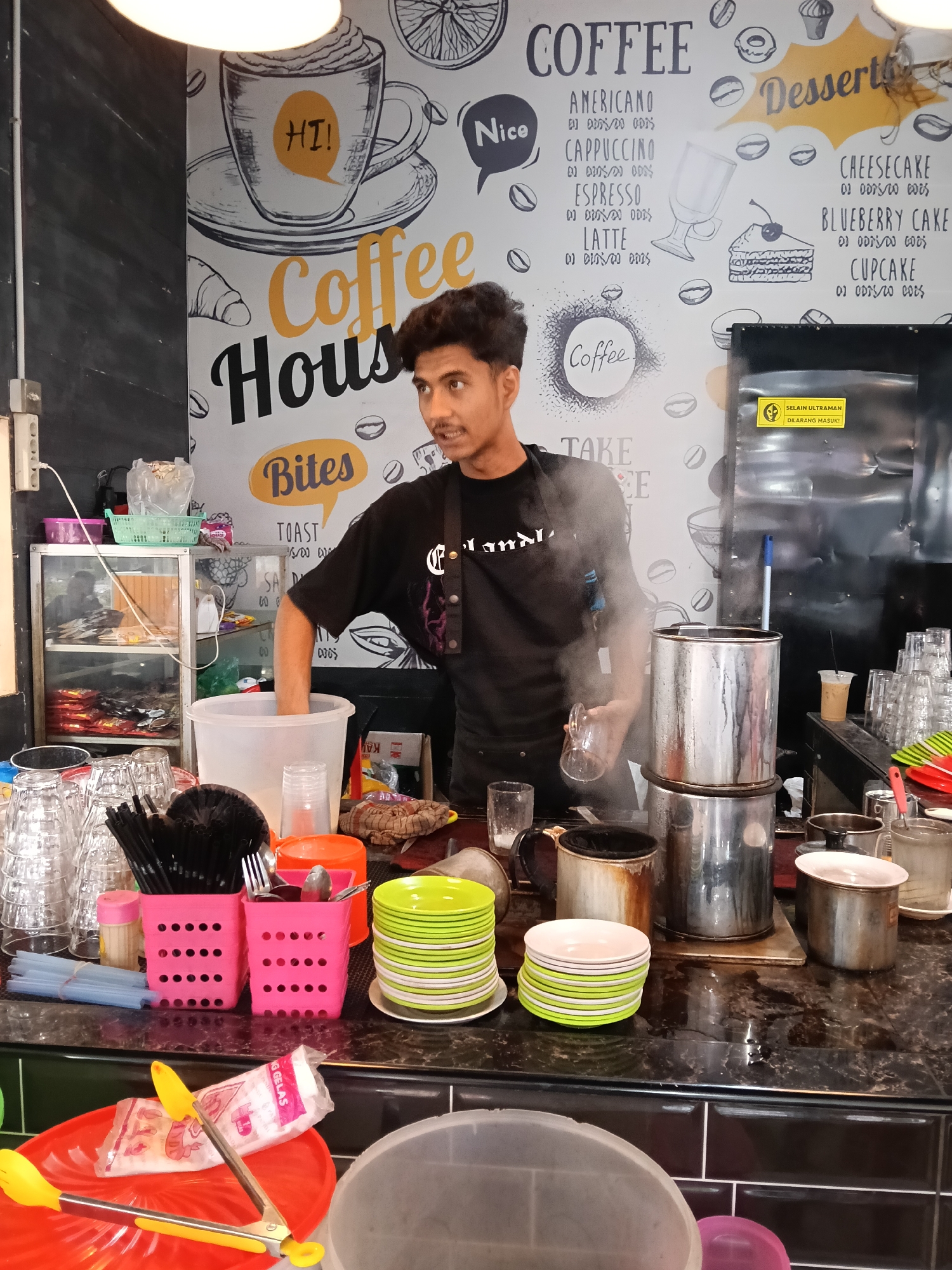 A young man stands behind a busy coffee shop counter in Lhokseumawe, Indonesia. He wears a black t-shirt and apron. Steam rises from metal coffee brewing equipment on the counter. The counter is cluttered with plates, cups, utensils, and containers. A wall mural behind the counter features coffee-related images and text, including a menu and the words "Coffee House Bites." A refrigerator is visible behind the man to the right.