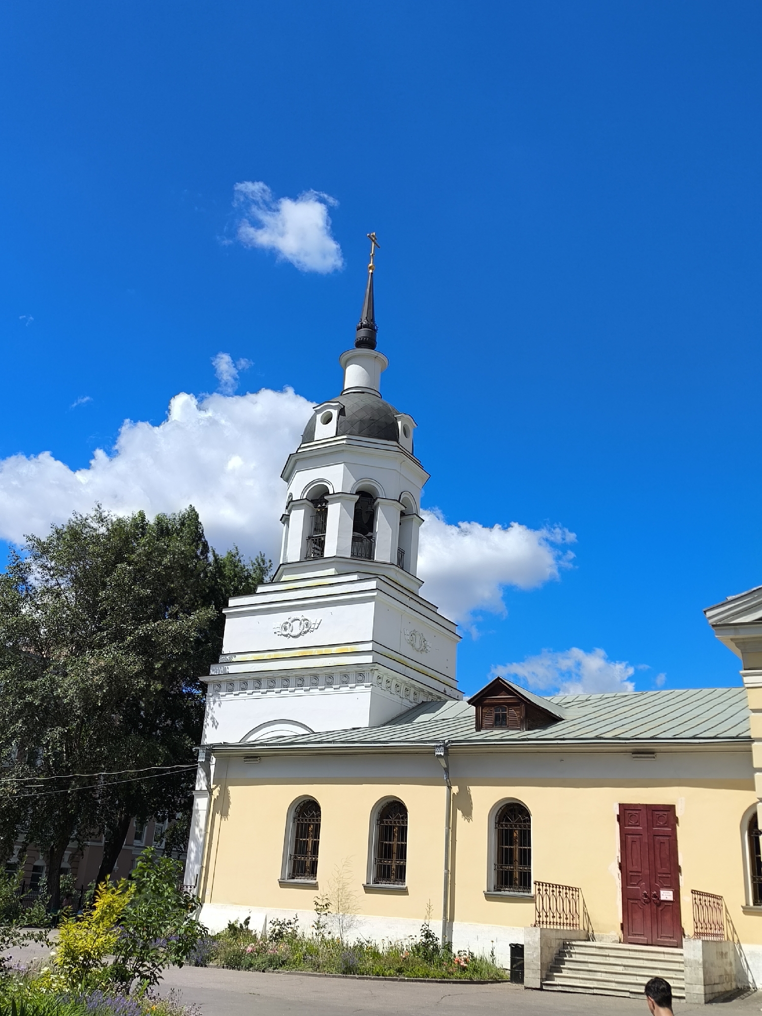 A low-angle shot shows the upper portion of a white and yellow church against a bright blue sky with scattered white clouds. The church features a white bell tower with a black dome and a golden cross at its apex. The main body of the church is painted a pale yellow and has arched windows with dark metal grates. A closed, dark red double door with a railing and stairs leads to the church entrance. To the left of the church, a large, leafy green tree is visible. The foreground shows a sidewalk or path with a variety of green plants and purple flowers. A person's head and shoulders are visible in the bottom right corner, facing away from the camera.