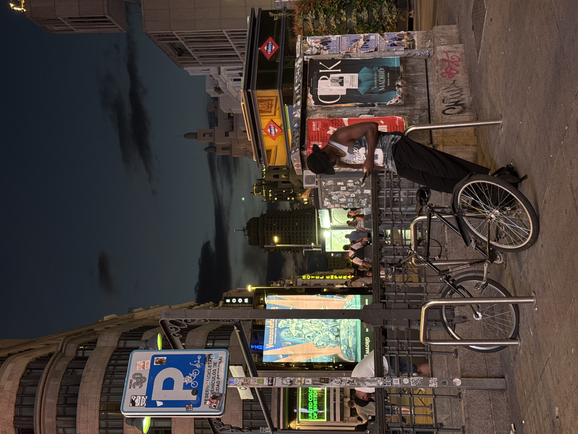 A young Black man wearing a bucket hat, a tank top, and baggy pants leans against a metal railing while looking at his phone. He is standing on a sidewalk in Madrid, Spain, at dusk. Behind him, a bicycle is parked in a rack. In the background, buildings and billboards line a street. A blue and white parking sign with a bicycle icon is mounted on a pole to the left. The sky is dark blue with some clouds.
