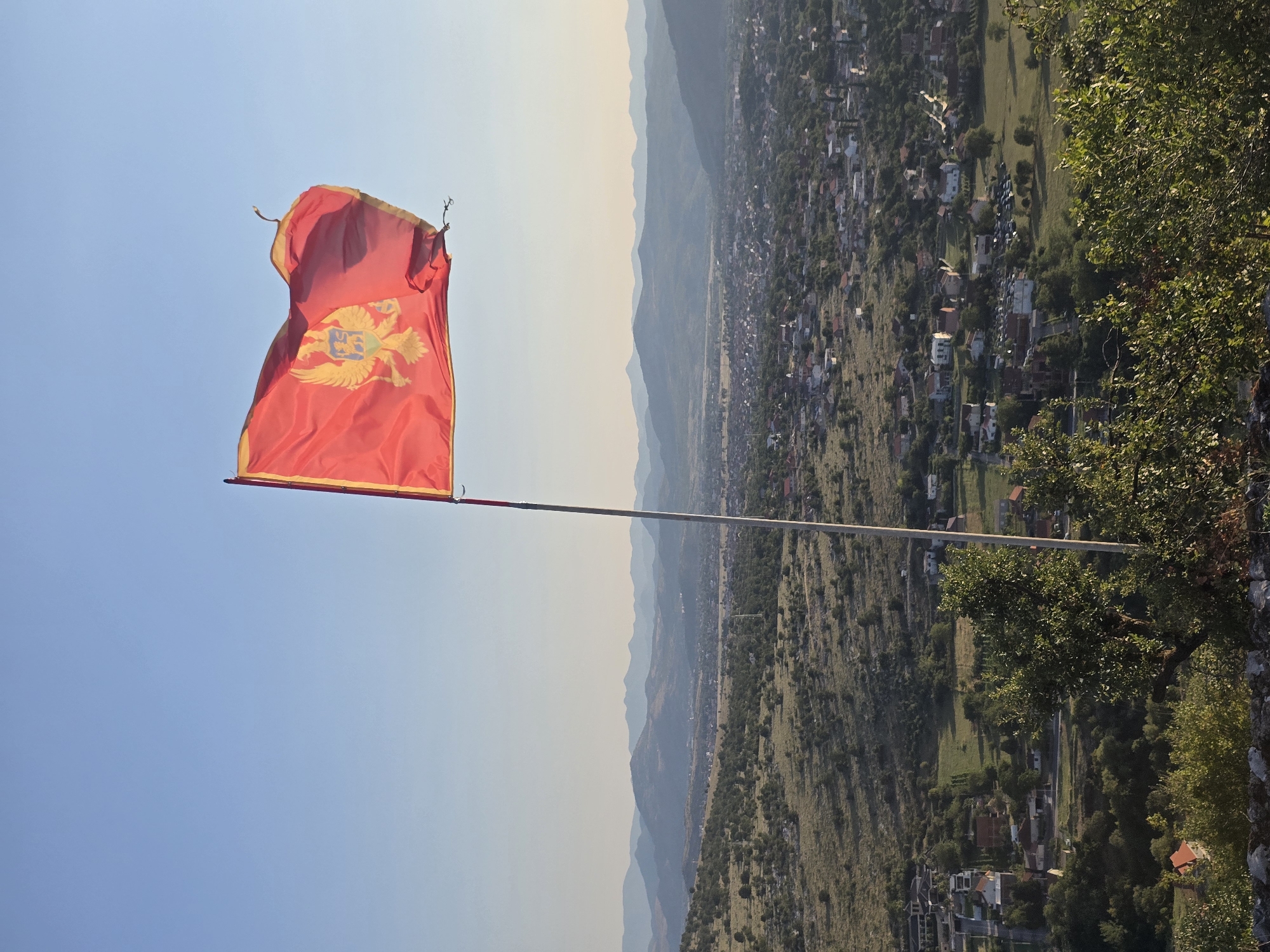 A red flag with a golden double-headed eagle emblem is flying from a pole against a clear blue sky. The flag is positioned on the right side of the frame, partially obscuring the background. Below the flag, a panoramic view of a valley unfolds, dotted with houses and trees. Distant, hazy mountains form the backdrop, their peaks fading into the horizon. The landscape appears to be a rural or semi-urban area, with a town or village nestled in the valley. The overall lighting suggests it is daytime, with the sun casting a warm glow over the scene.