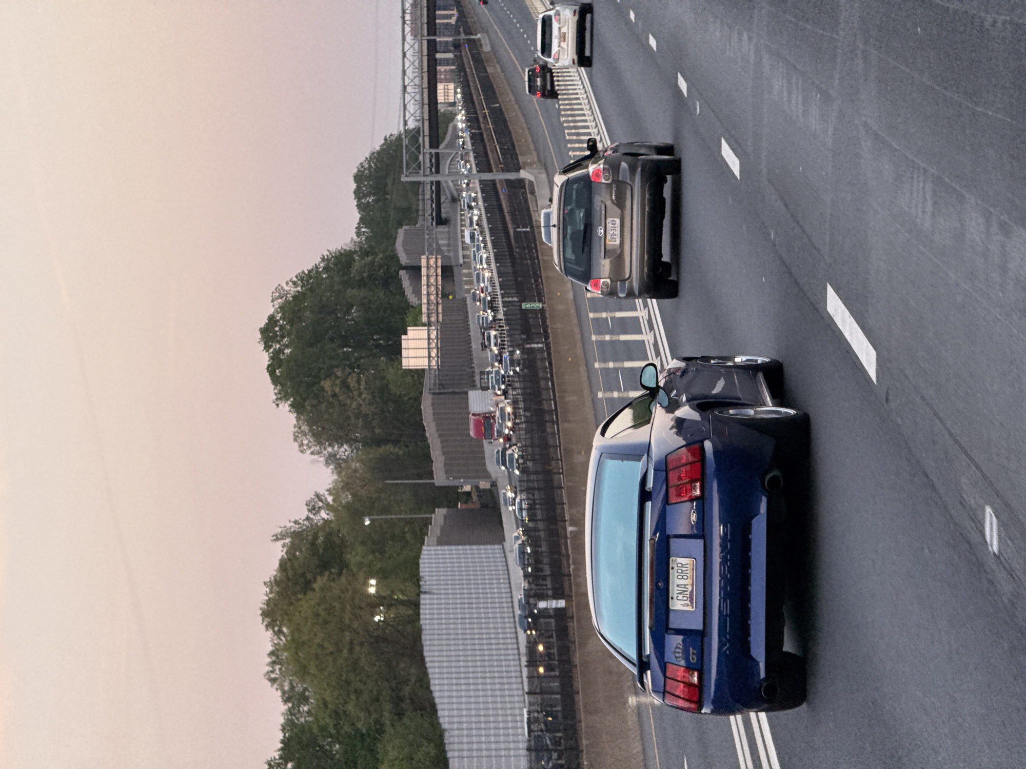 The image, taken from an elevated perspective, captures a multi-lane highway and an adjacent railway line, likely within Vienna, Virginia, United States. The time of day appears to be late afternoon or early evening, evidenced by the soft, diffused light and the pale, warm-toned sky. The weather is clear.
In the foreground, a blue Ford Mustang GT is prominently featured, facing away from the viewer in one of the active highway lanes. Its Virginia license plate is clearly visible, displaying the alphanumeric sequence "GNA 8RR." The "GT" emblem is also seen on its rear. Ahead of the Mustang, in the same lane, is a gray SUV or crossover, with its license plate reading "TFD-3149."
To the left of these vehicles, separated by a median and a railway track, is another section of the highway experiencing significant congestion, with a long line of vehicles appearing slow-moving or stopped. The railway line itself runs parallel to the highway. Trees dense with foliage line the sides of the roads and railway, with some buildings partially visible through the greenery. Overhead, parts of a bridge structure or power lines can be seen spanning the area. The scene depicts typical highway activity, contrasting flowing traffic with heavily congested lanes.