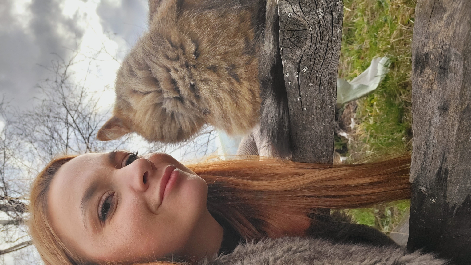 The image is a selfie taken outdoors in Зипуново, Russia, capturing a young woman and a cat in a close-up, low-angle perspective. The woman, positioned in the foreground, has fair skin, light make-up, and long, reddish-brown hair cascading around her. She looks upwards and slightly to the right with a soft, gentle smile, her eyes conveying a calm expression. She appears to be wearing a dark garment with a textured, possibly faux fur, collar that matches the greyish fur visible around her shoulder.
Above and to the right of the woman, a fluffy, tabby-patterned cat, with fur in shades of brown, grey, and cream, is partially visible. Its head is angled downwards, as if it is looking at or interacting with the woman, though its full face is not shown, only a portion of its profile and one ear.
The background consists of a bright, overcast sky with bare tree branches silhouetted against it, indicating a season like late autumn, winter, or early spring. To the far right, two weathered wooden fence posts or planks stand vertically, showing pronounced grain, cracks, and patches of lichen. Between these wooden elements, patches of green grass and what appears to be a discarded white plastic bag or sheet are visible on the ground.
The scene depicts a peaceful, candid moment of interaction between a woman and her pet on a cloudy daytime, outdoor setting. There is no visible text in the image.