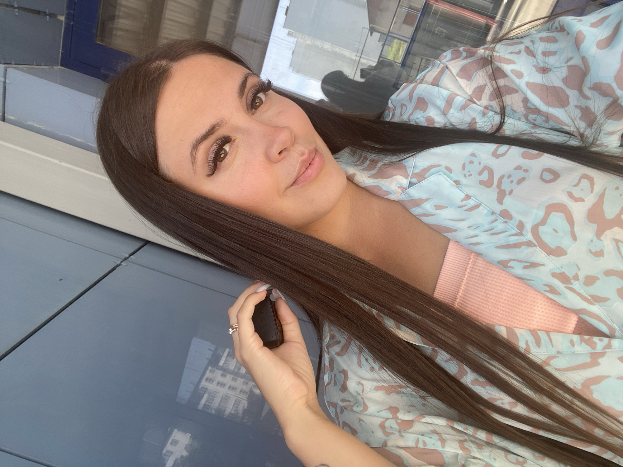 In a selfie taken outdoors, a woman with long, dark brown hair is positioned in the foreground. She is wearing a pink ribbed tank top underneath a light blue and pink leopard-print open robe. Her right hand is raised to her head, holding a small black object, and she wears two rings on her right ring finger. Her makeup is visible, with emphasis on her eyes and lips. In the background, a modern building with reflective blue panels and windows is visible, with some lighter colored buildings reflected in the glass. The lighting suggests it is daytime. The specific location of Chaikovsky, Russia, is not ascertainable from the image itself, as there are no distinguishing landmarks or contextual clues within the photograph that would indicate its geographical origin.