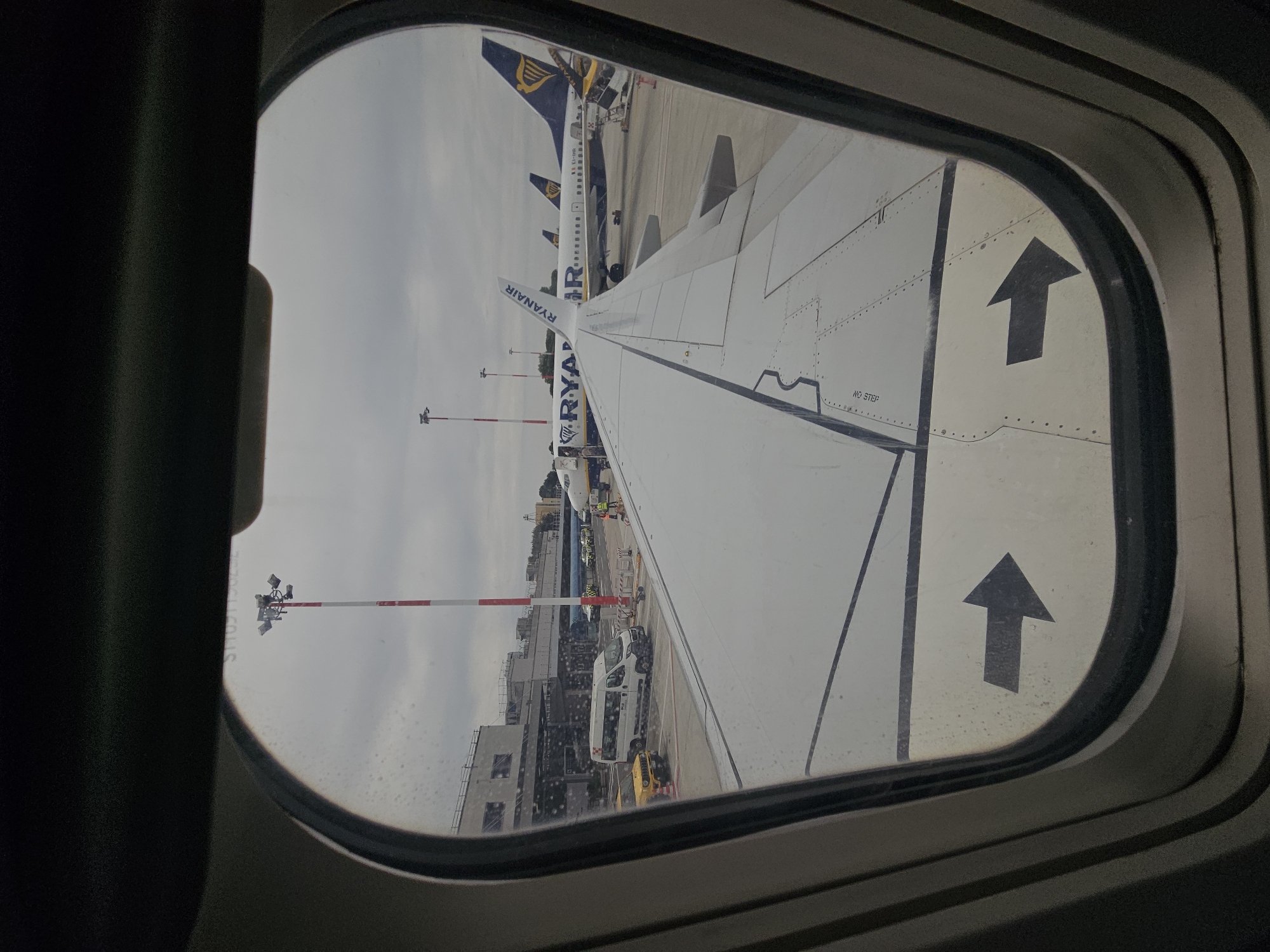 The image shows an airplane wing as seen through an airplane window. The plane is a Ryanair plane. Another Ryanair plane is visible in the background. The plane is on the ground near an airport building. There is a variety of airport ground vehicles, including a white van. There are red and white striped poles on the ground. The location is likely Rome, Italy, due to Ryanair's extensive presence at Rome airports.