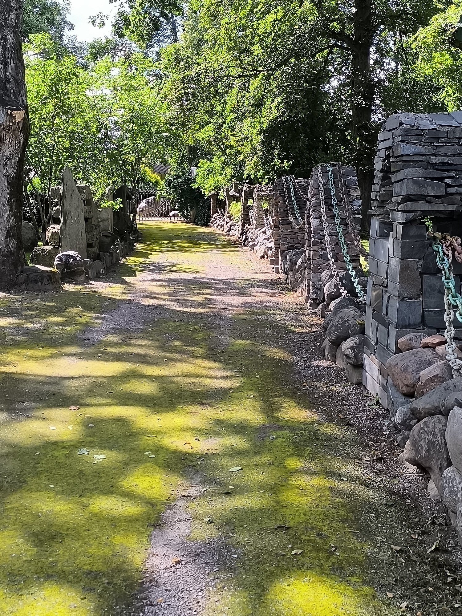 The image depicts a pathway leading through a park-like setting in the city of Mosėdis, Lithuania. The path, composed of gravel and patches of bright green moss, is flanked by stone structures. On the right side of the path, a wall constructed from stacked flat stones rises, adorned with several lengths of thick metal chain, some green and some silver. Large, rounded stones are integrated into the base of this wall. On the left side, the pathway is bordered by a similar arrangement of stone pillars and rocks, with some larger, more irregularly shaped standing stones visible. Trees with dense green foliage form a canopy overhead, casting dappled shadows onto the path. In the distance, a wrought-iron gate can be seen, suggesting an entrance to another area. The overall impression is one of a rustic, natural, and somewhat artful outdoor space.
