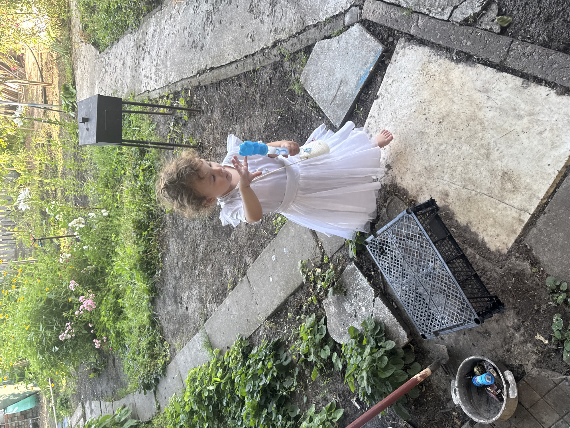 A young child, appearing to be a toddler, stands on a paved area in a garden. The child is wearing a white dress and holding a blue toy in their right hand. The child's hair is curly and light brown, and they are looking down at the toy. The ground is a mix of dirt and concrete slabs, with some greenery and plants growing around. In the background, there is a black metal stand with a square top, and a grey plastic crate lies on its side. A bucket filled with various items, including what appears to be cigarette packs and a blue bottle, is visible in the lower right corner. The scene appears to be outdoors, likely in a backyard or garden, with evidence of some disarray. The lighting suggests it is daytime, possibly late afternoon due to the shadows. The overall impression is of a child playing in a somewhat unkempt outdoor space. There is visible text on the cigarette packs in the bucket, which appears to be "KENT". The location context is Shakhty, Russia.
