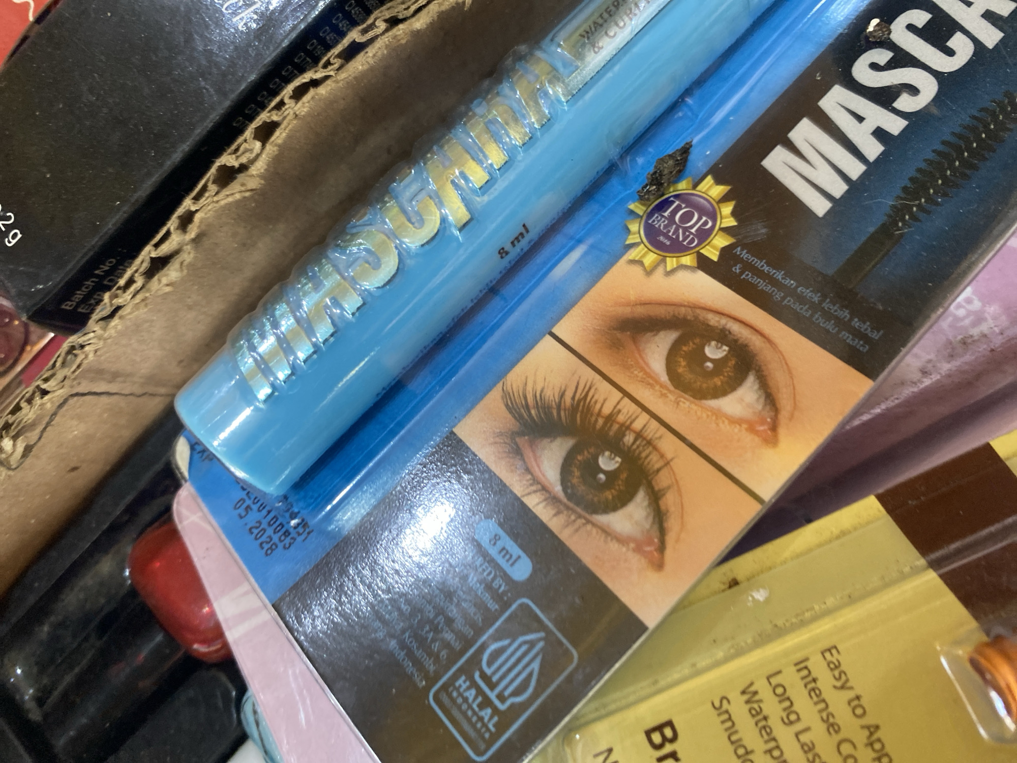 The image shows a close-up view of a box of mascara. The box is blue and white with the word "MASCARA" printed in large white letters. There is also an image of an eye on the box. The mascara tube itself is blue and has the word "MASCARA" printed on it in gold letters. The image appears to be taken in a store or a home setting, with other items visible in the background, including what looks like makeup and a cardboard box. No specific location in Ta', Indonesia can be determined from the image.