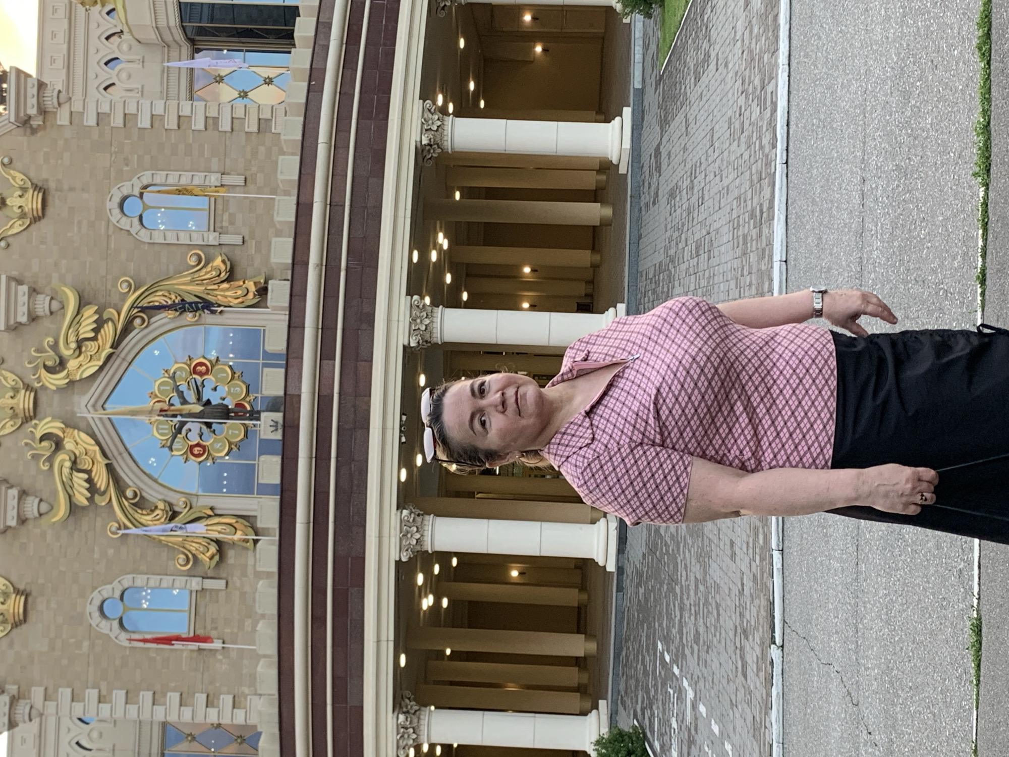 A middle-aged woman with blonde hair, wearing a pink checkered shirt and a black skirt, stands in front of a building with classical columns. She is wearing a watch on her left wrist and a ring on her right hand. The building is ornate, with gold accents and large windows. The ground in front of the building is paved with bricks. The woman is looking at the camera. The scene appears to be in the city of Kazan, Russia, likely in front of the "Miracle of the Clock" at the Kazan Family Center.